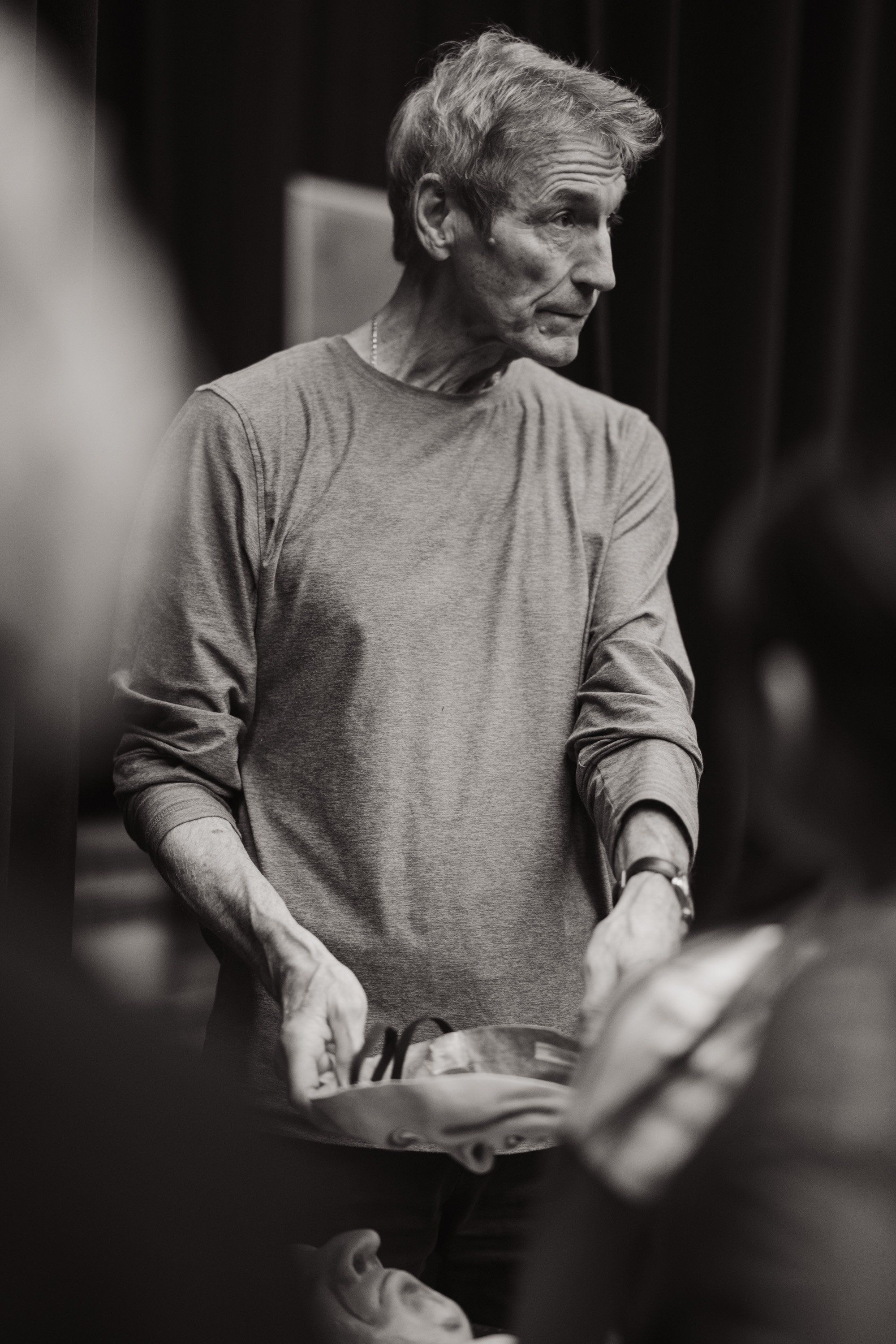 Older man in grey long-sleeved shirt speaks thoughtfully during performance, captured in black and white against dark background