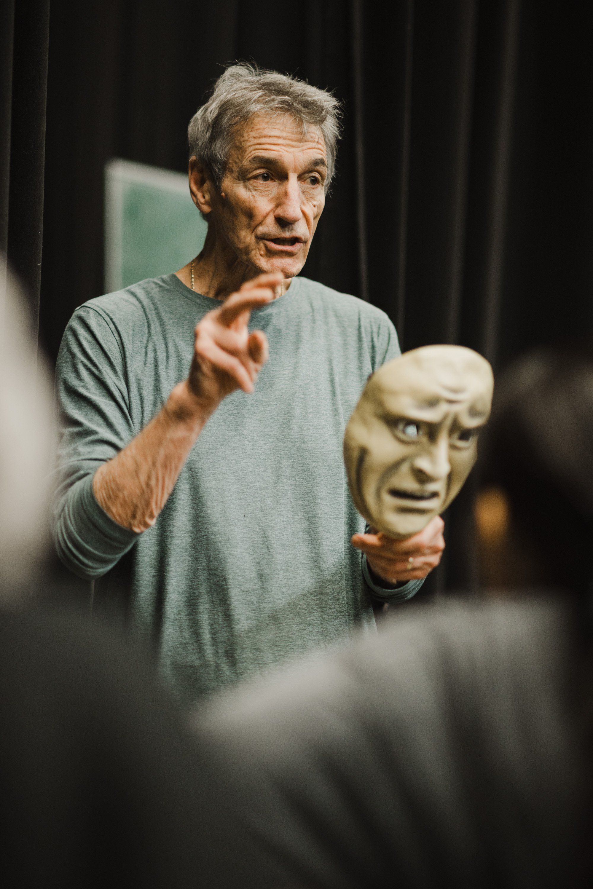 Artist gestures expressively while holding a carved mask, speaking to an audience in an intimate theater setting