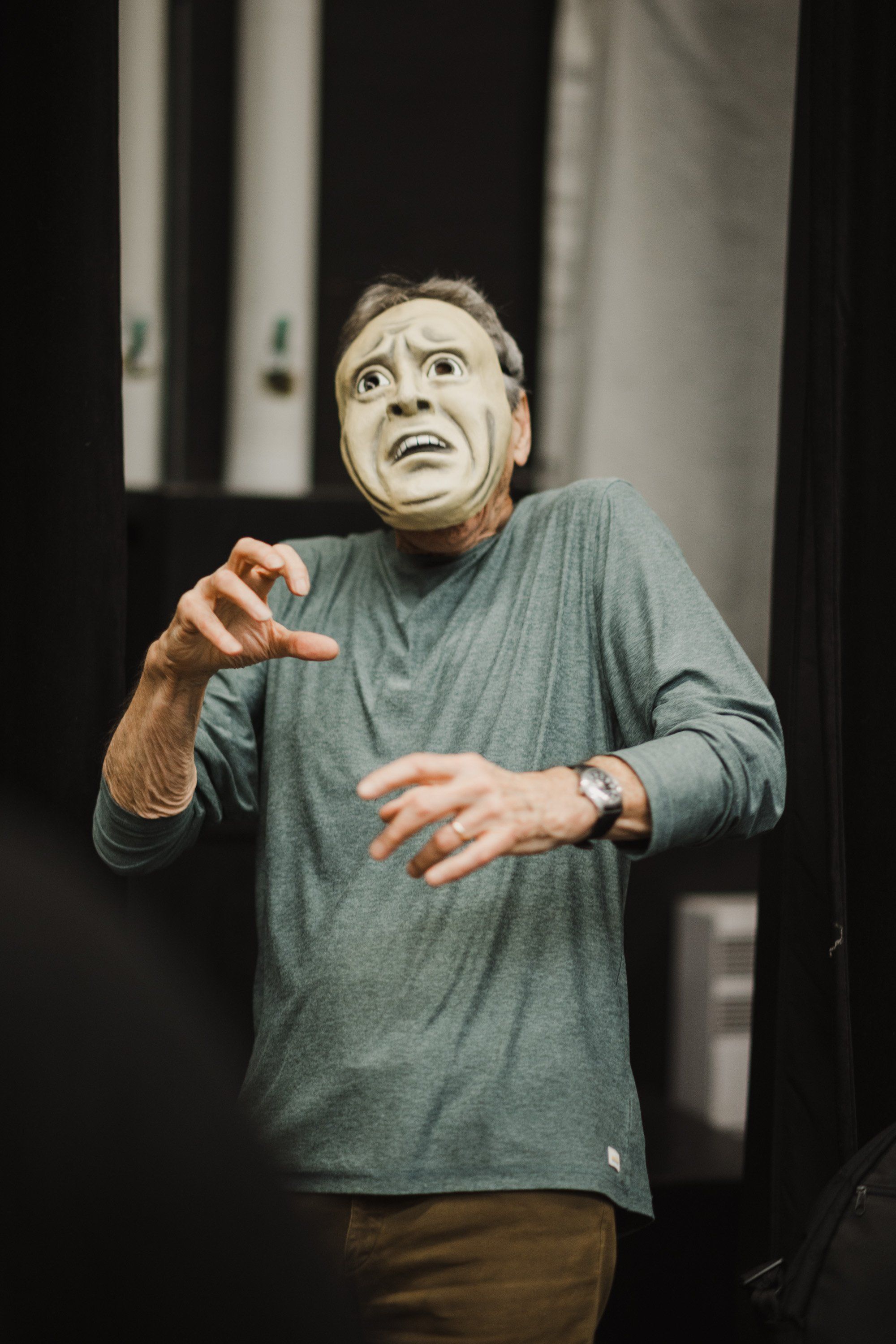 Performer wearing pale mask gestures expressively on stage in gray-green shirt, dramatic lighting highlighting emotional intensity