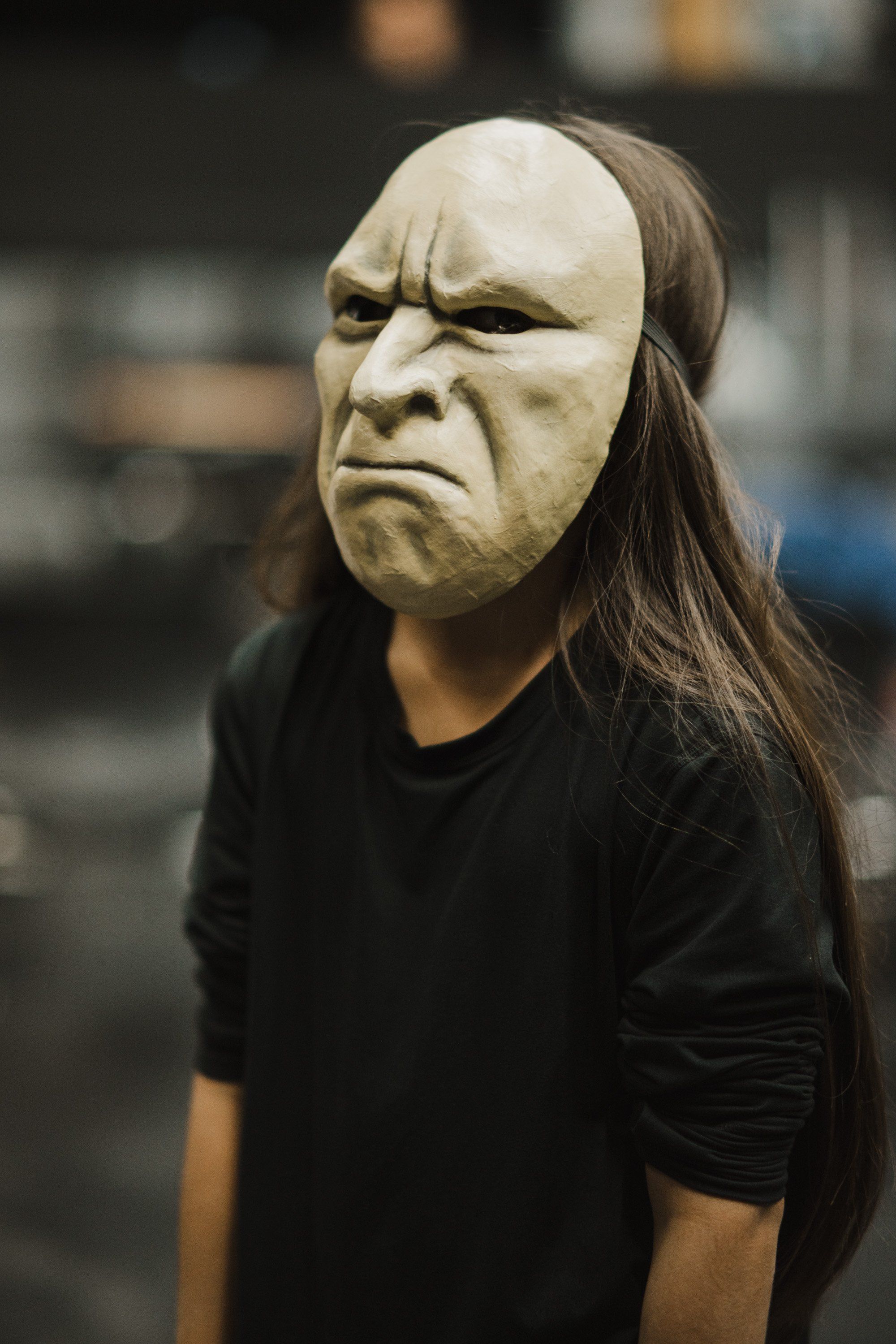 Person wearing long dark hair and pale mask with stern expression, black t-shirt, indoor blurred background