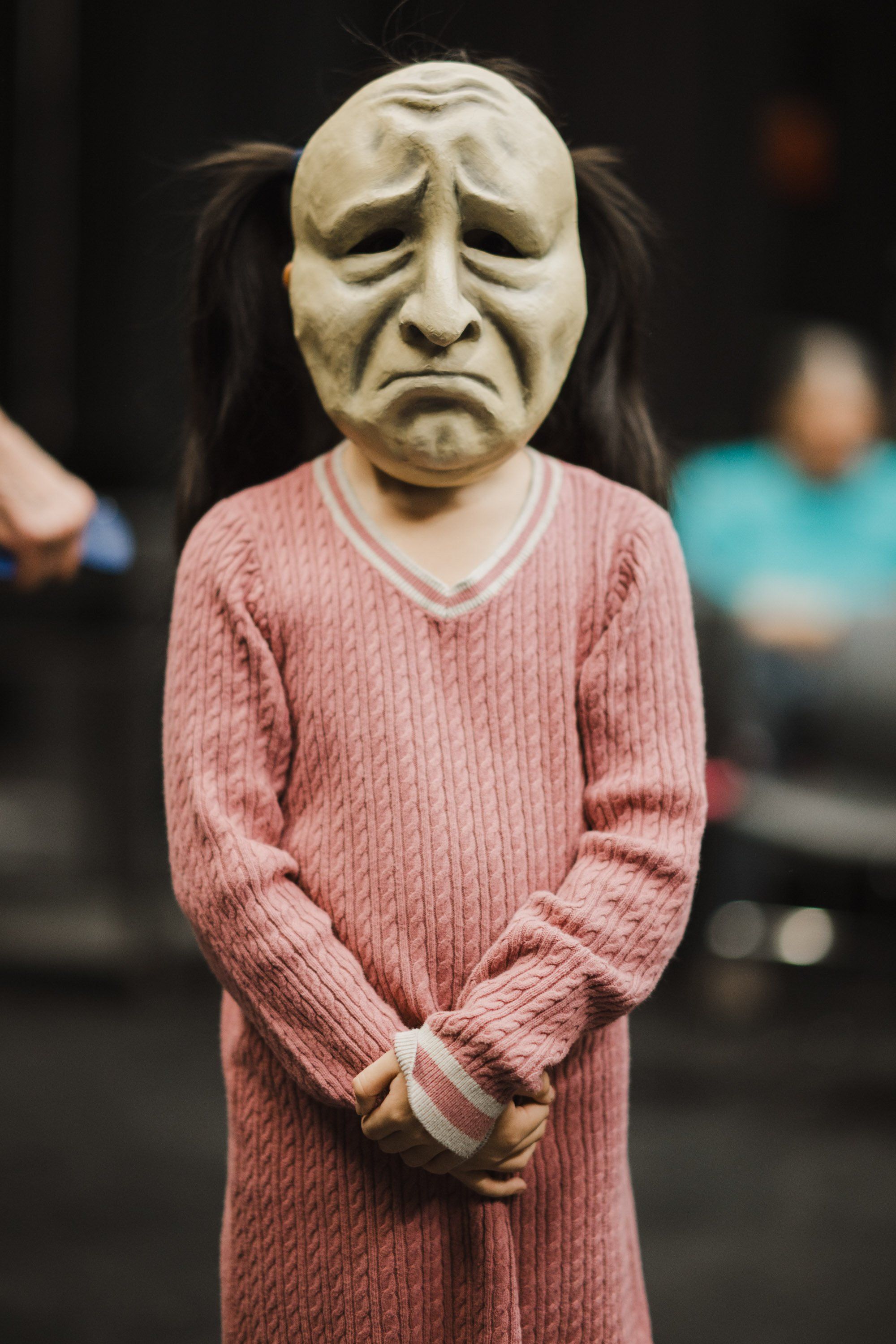 Figure in carved wooden mask and pink ribbed sweater with hands clasped, performing at indoor cultural event