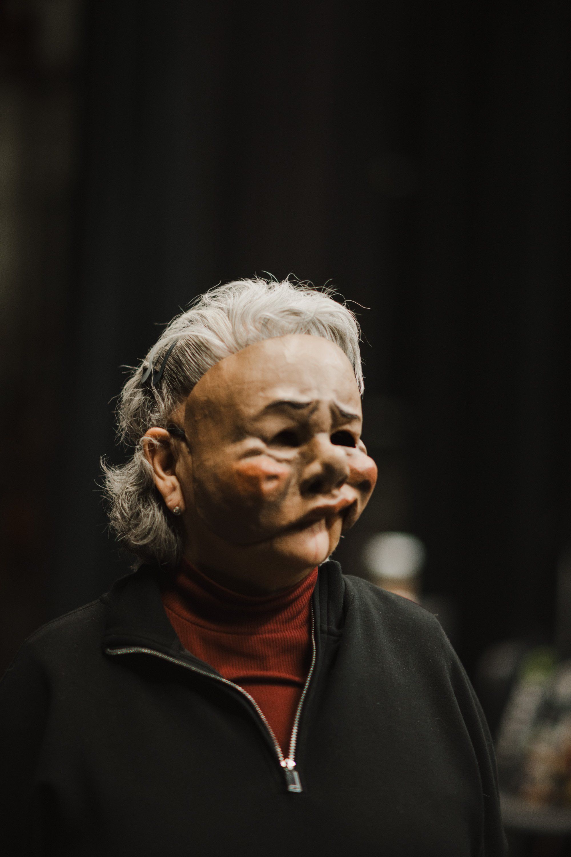 Older woman with white hair wearing a red turtleneck and black jacket, photographed against a dark background with warm side lighting