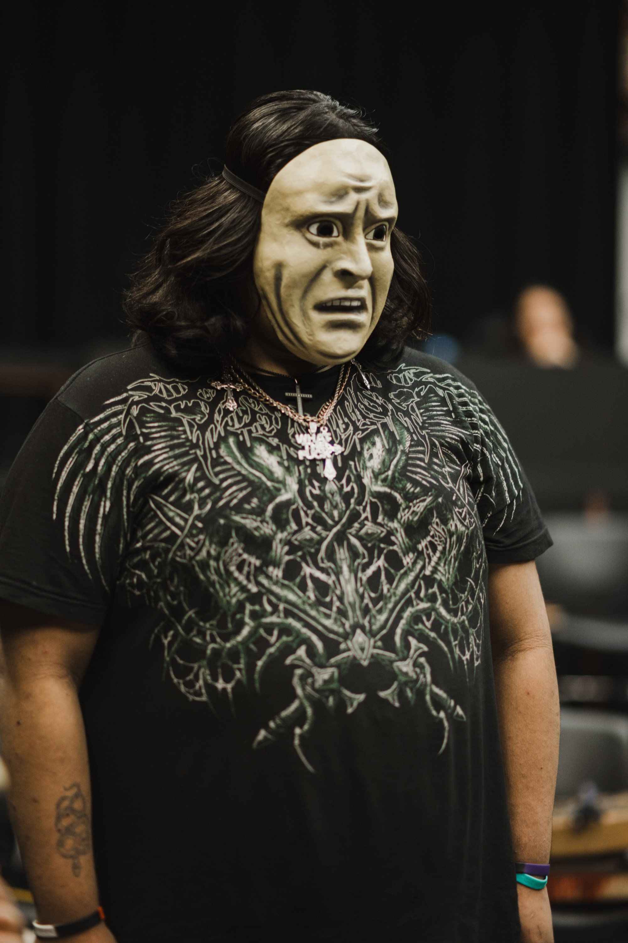 Performer in pale mask with long dark hair wears intricate black graphic shirt against dark background, intense gaze directed forward