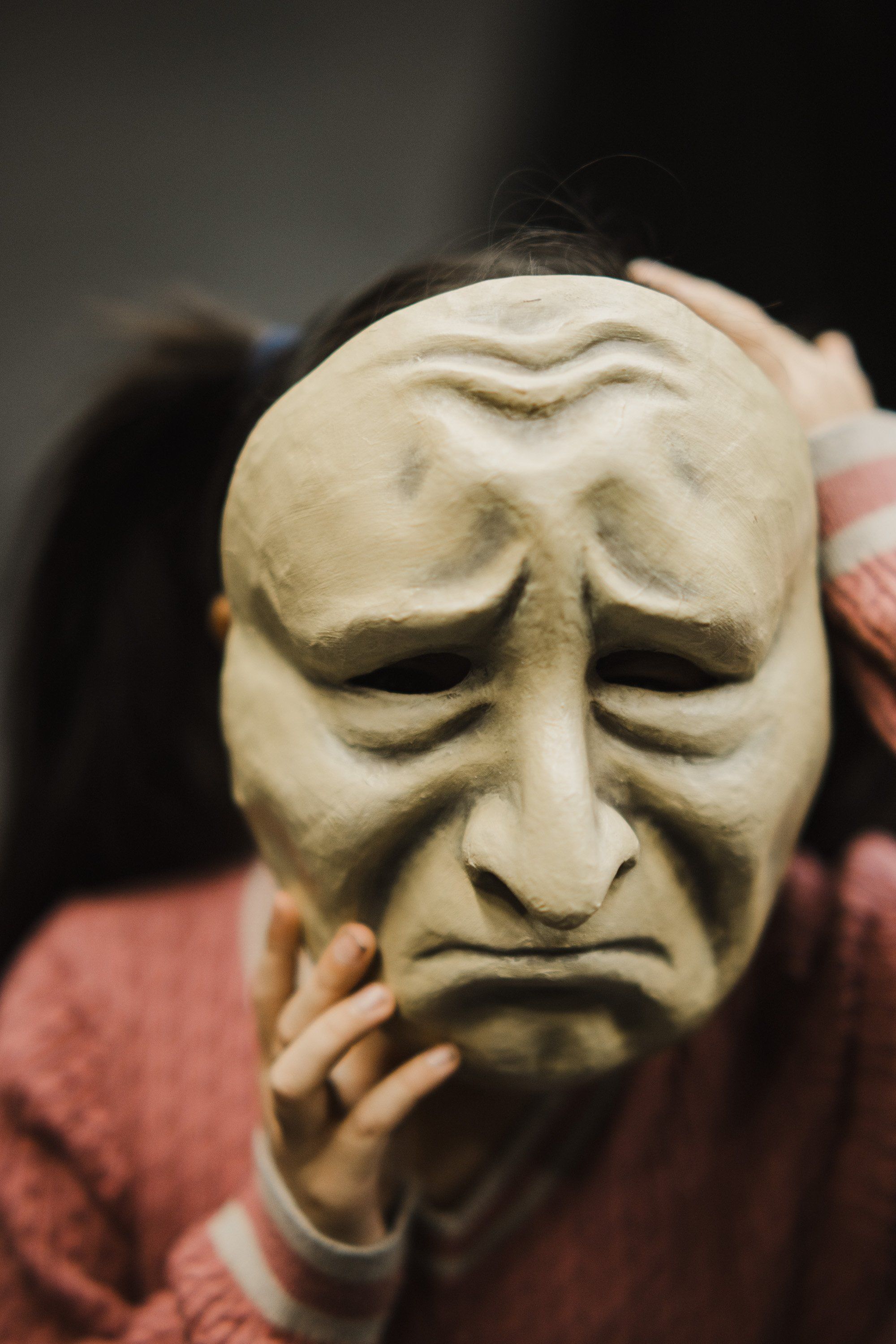 Carved wooden mask with closed eyes and sorrowful expression, held in hands wearing coral-colored sleeves against dark background