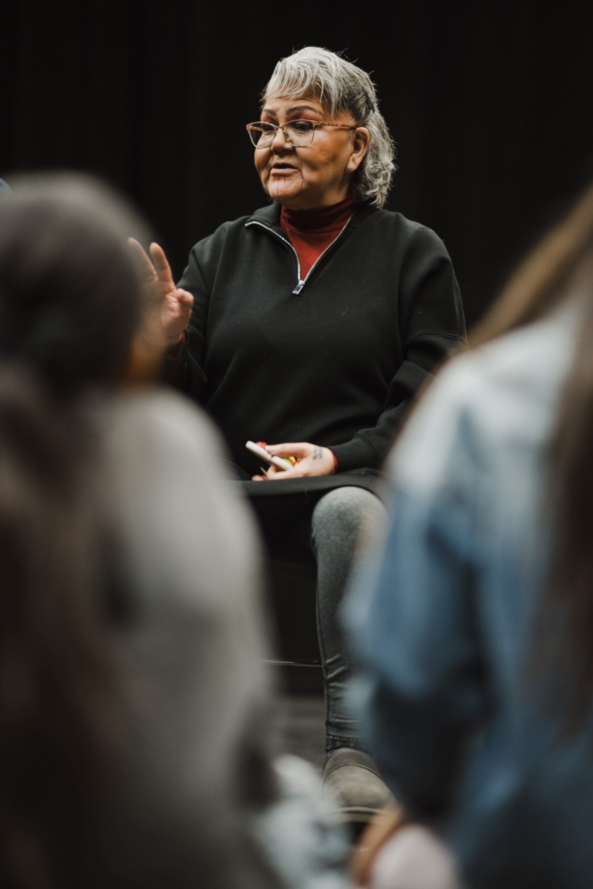 Woman with white hair and glasses in black jacket speaks to seated audience members during Coast Salish cultural event