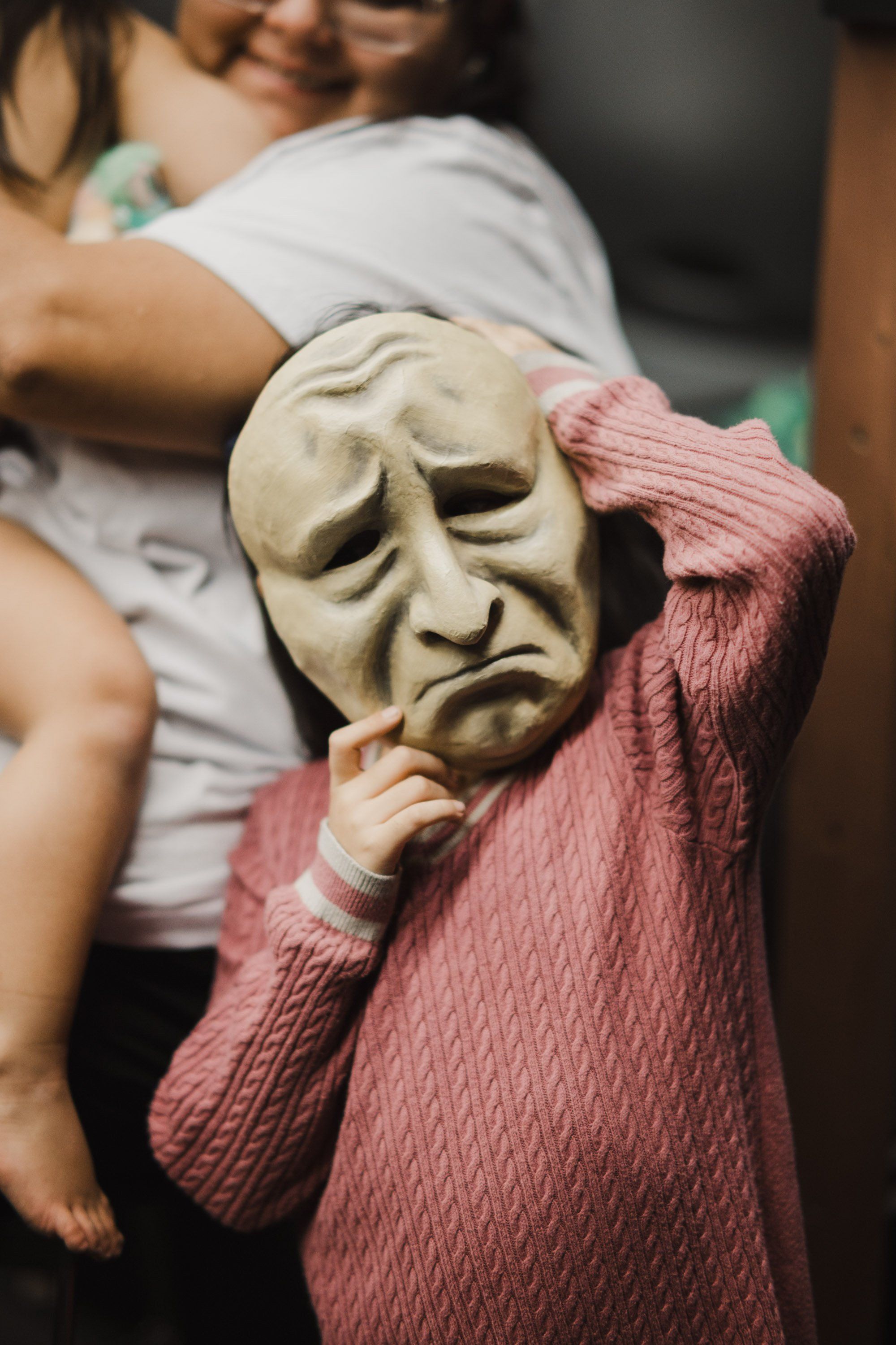 Child in pink cardigan holds carved wooden mask to face while adult holds them, both backlit indoors
