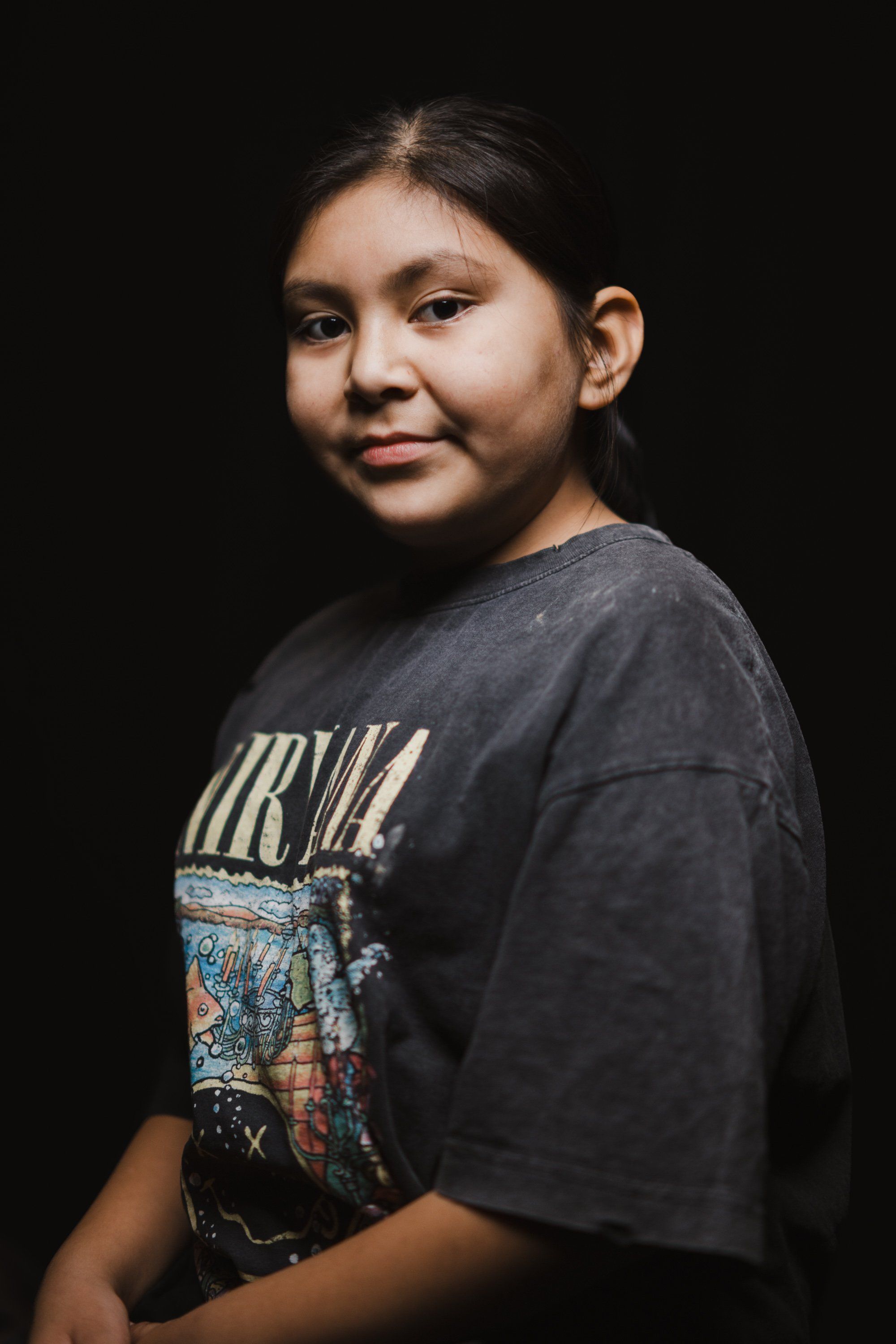 Young person in a dark band graphic t-shirt turns toward camera against black background, warm studio lighting illuminating their face and gentle expression