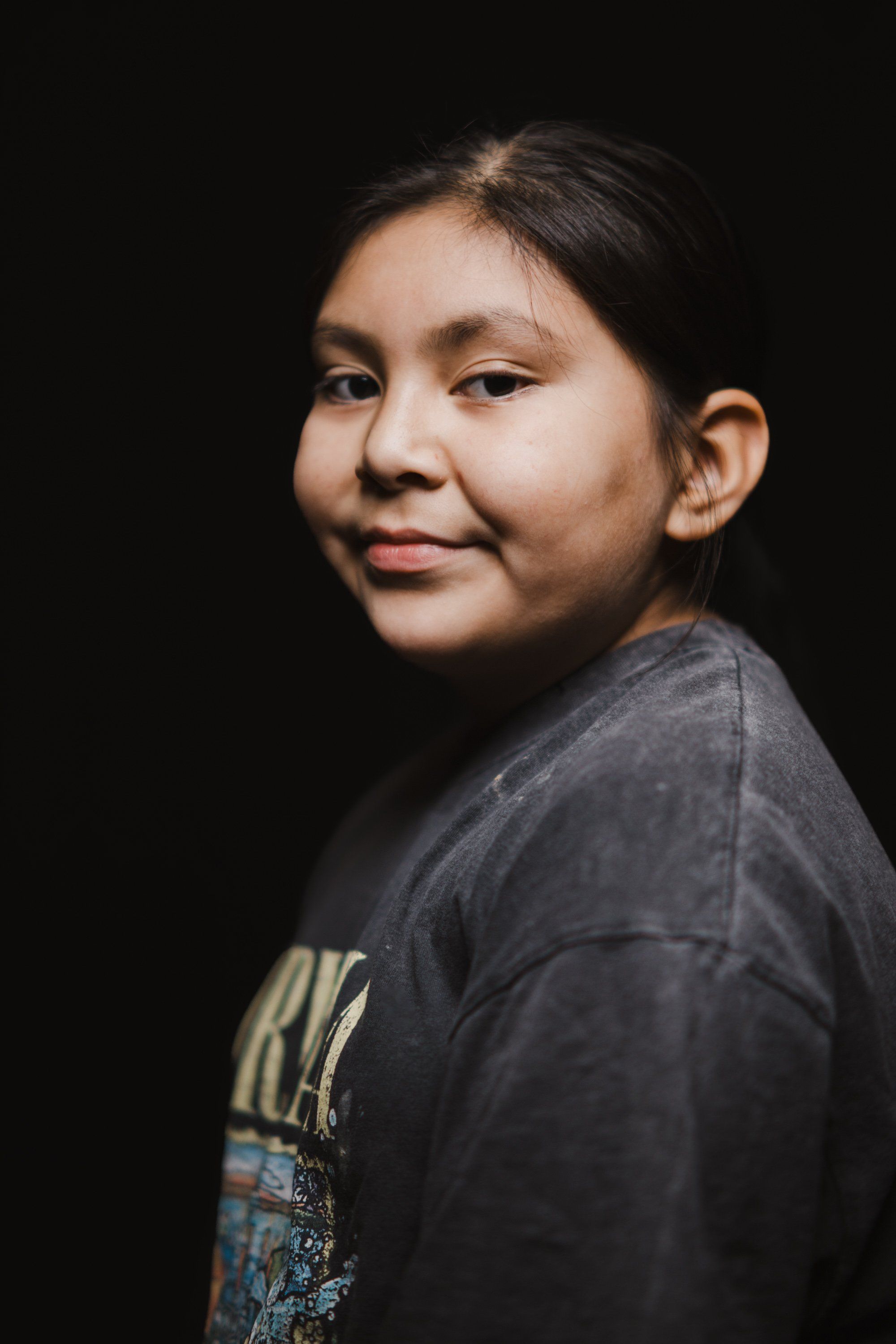 Young person in dark graphic t-shirt gazes over shoulder against black background, warm studio lighting highlighting their face