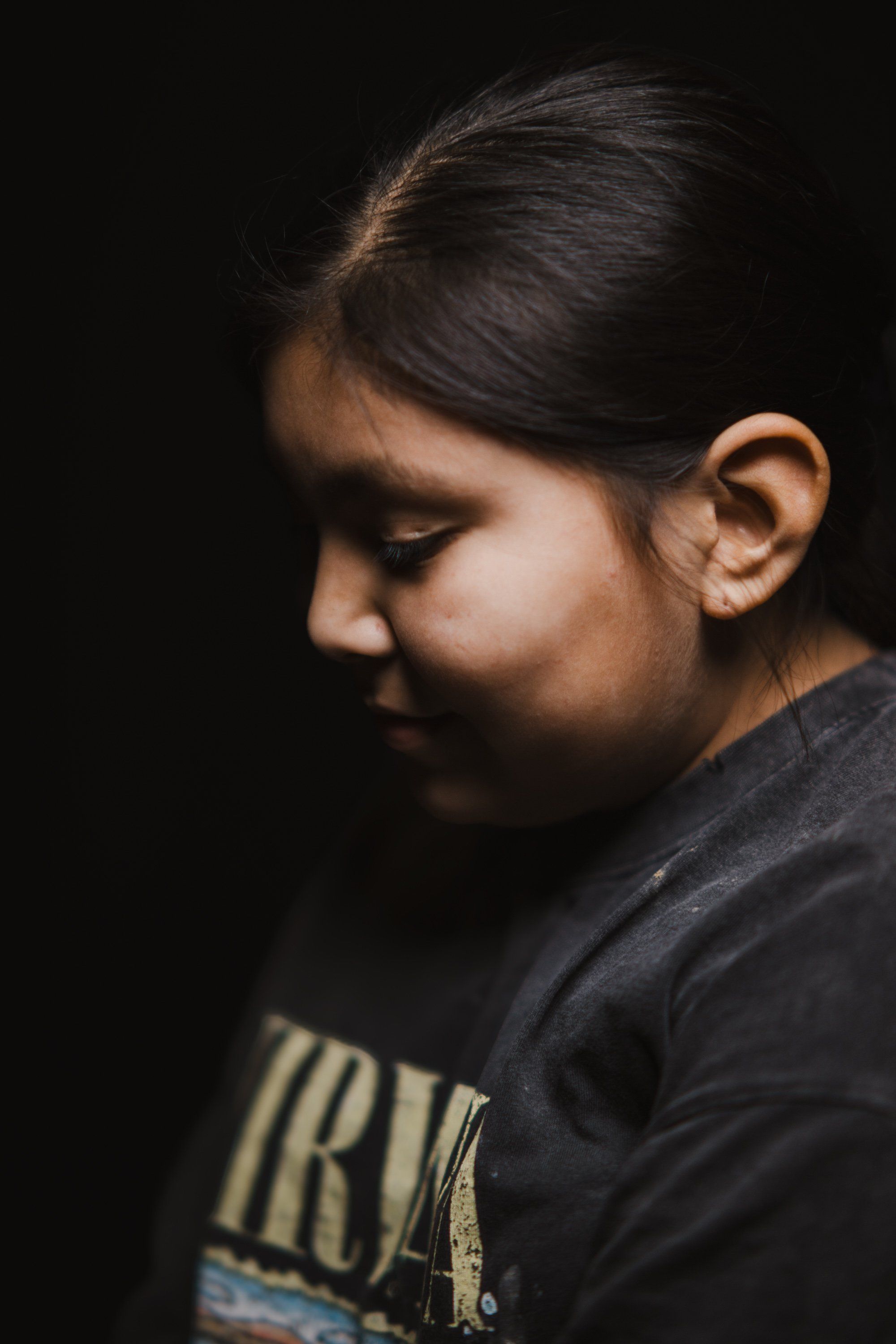 Young person in profile against dark background, wearing black graphic tee, head bowed in contemplative moment with warm side lighting