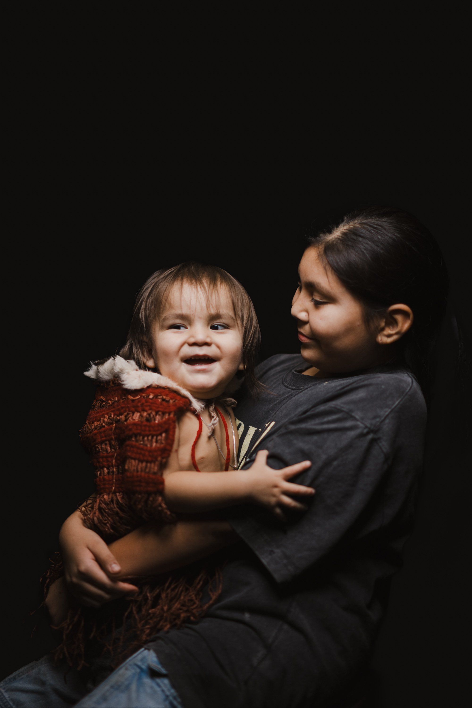 Older child holding smiling toddler against dark background, both wearing earth-toned clothing, intimate family portrait