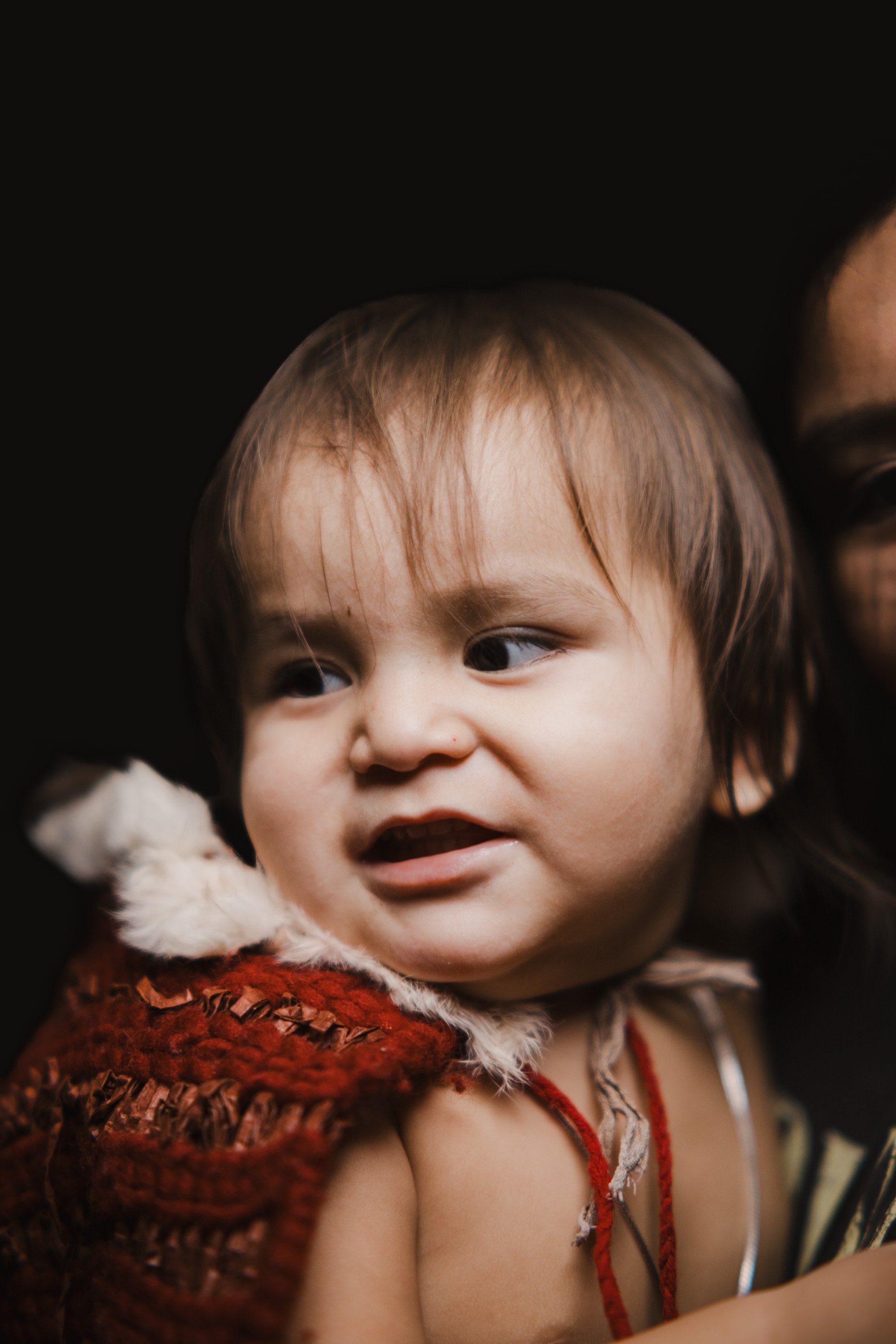 Toddler in russet-red regalia with white fur trim smiles warmly against a dark background, held by an adult partially visible at frame's edge