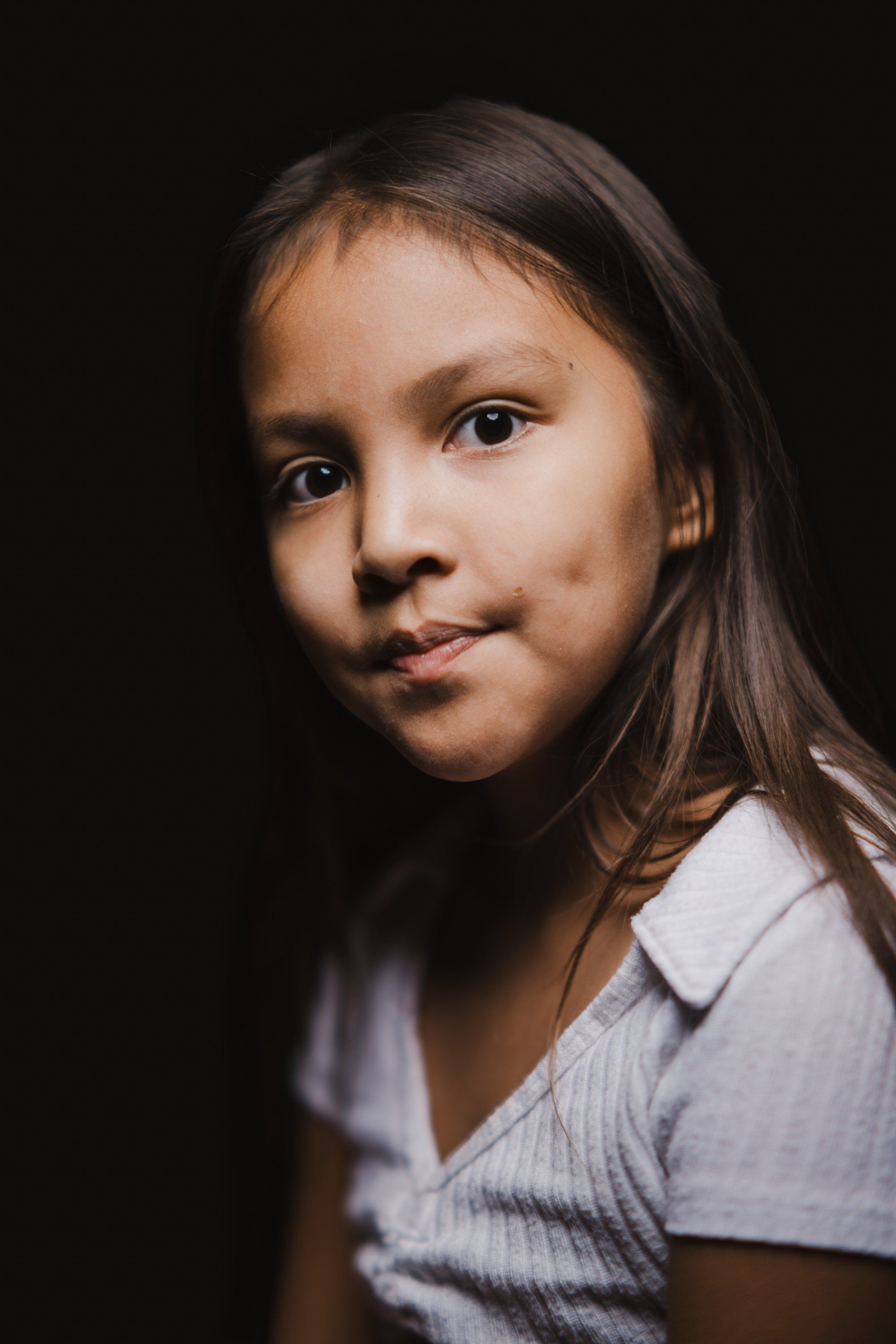 Young girl with long dark hair in white shirt against black background, gentle side-lit portrait with warm, contemplative expression