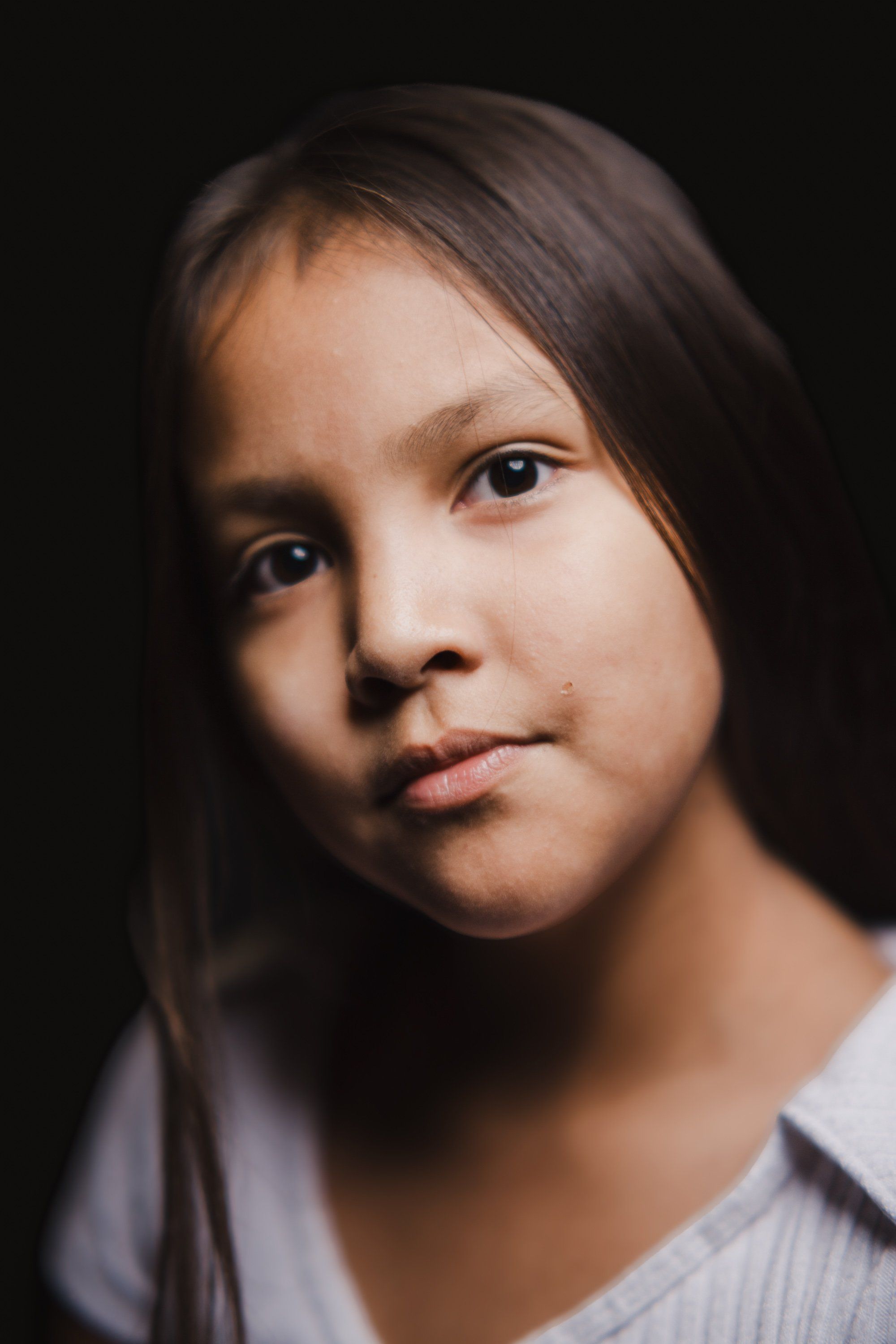 Young person with long dark hair gazes thoughtfully at camera against black background, softly lit portrait