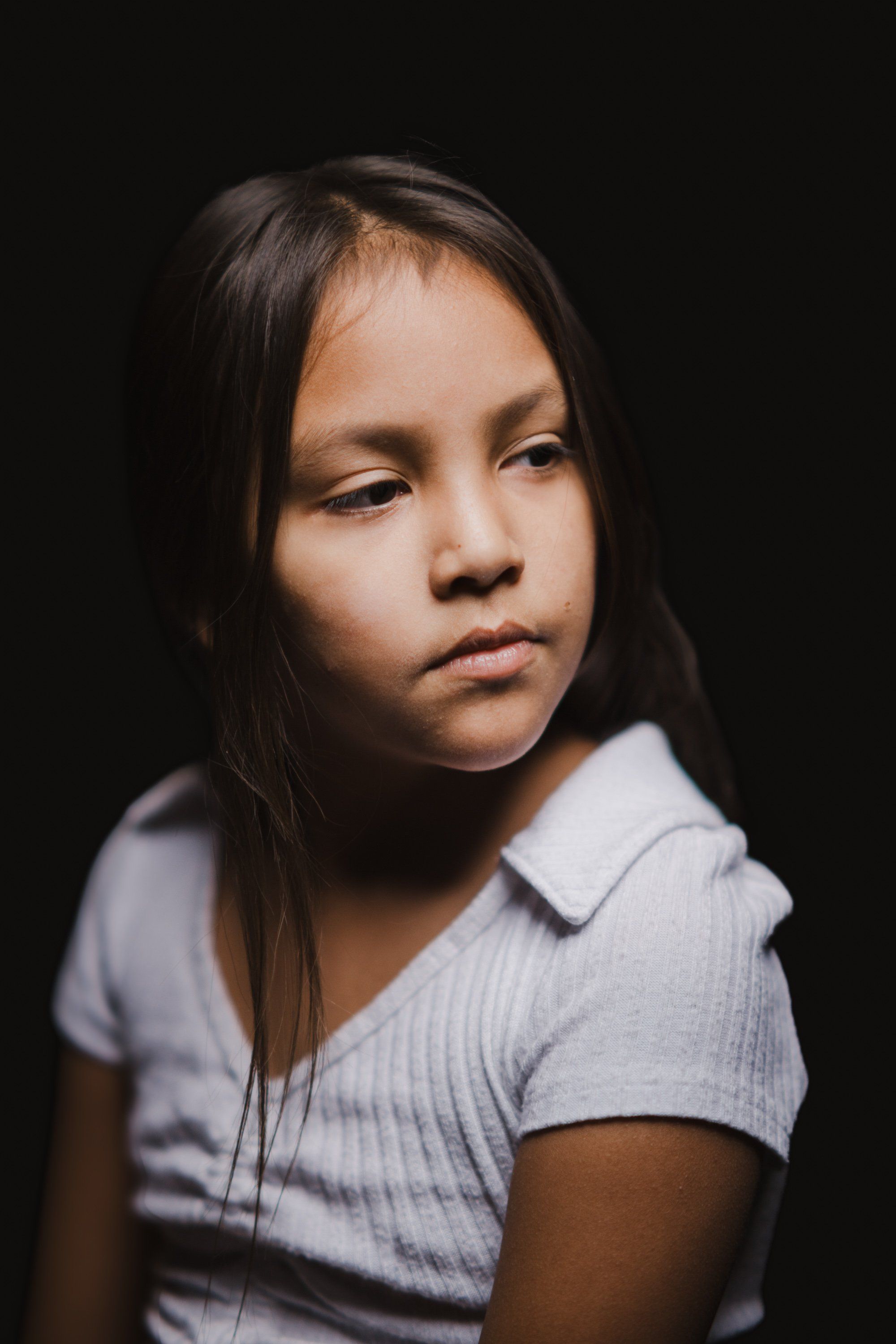 Young person with long dark hair wearing a light gray ribbed shirt against a black background, gazing downward with a thoughtful, serene expression