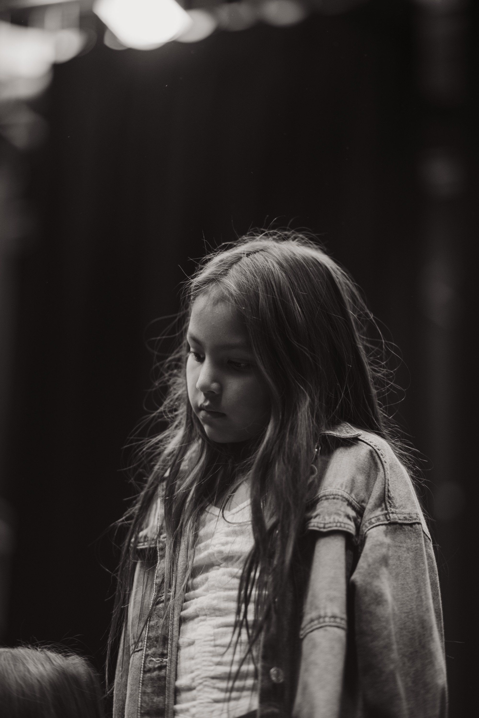 Young girl in denim shirt gazes thoughtfully to the side, backlit against dark background in black and white portrait