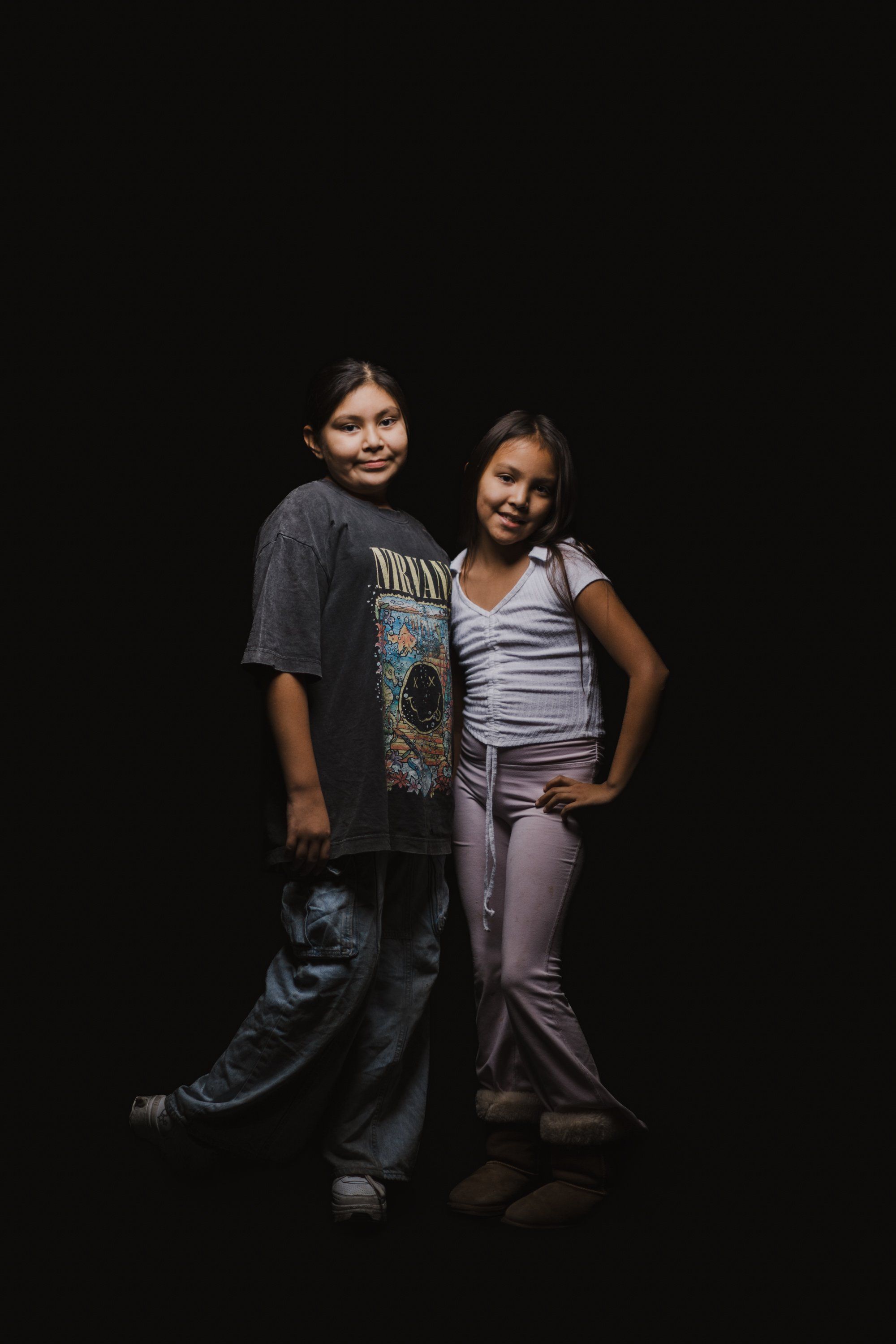 Two young people stand together against a black background, warmly posed with arms around each other, wearing casual clothing in soft studio lighting