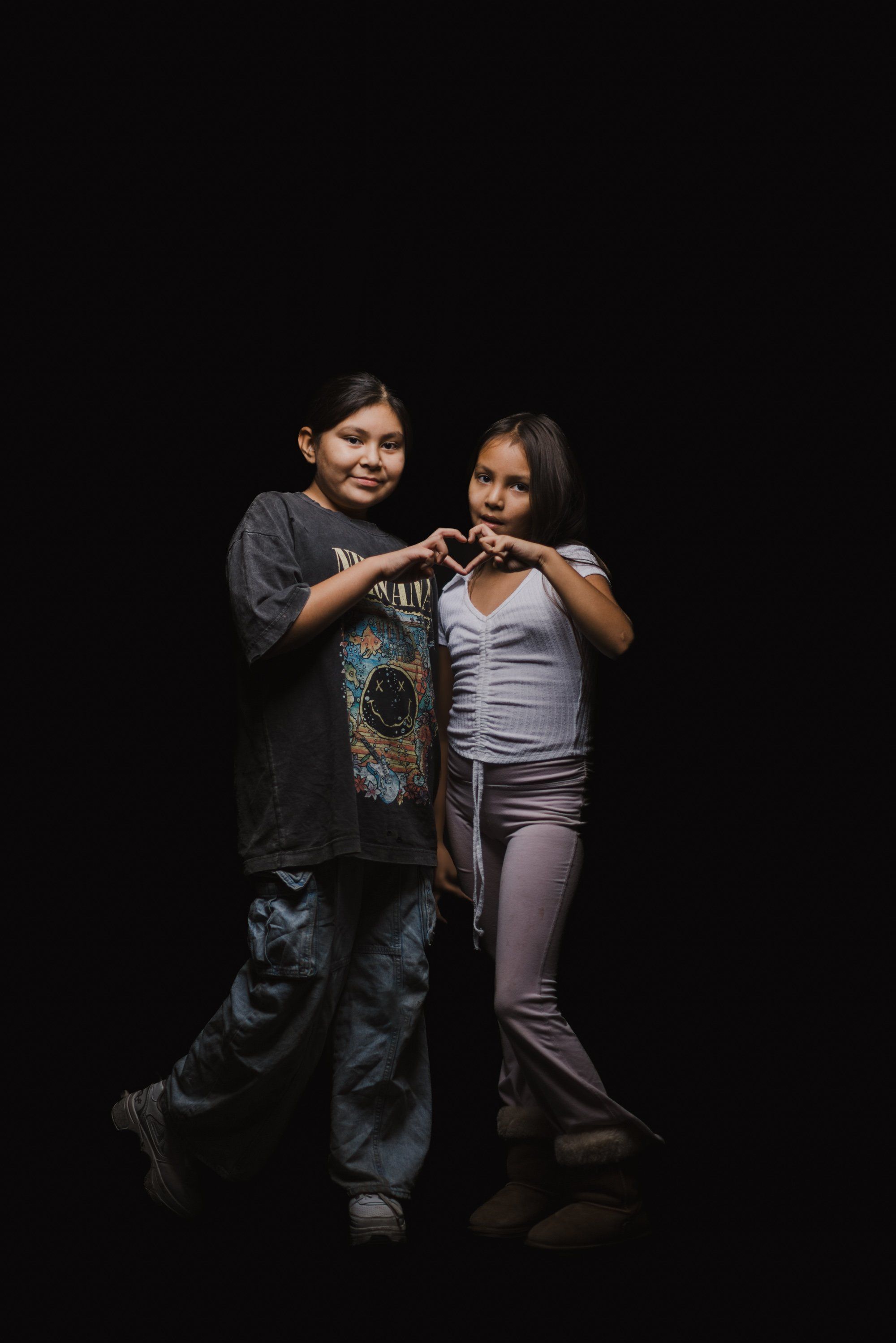 Two young people form a heart shape with their hands against a black background, smiling warmly at the camera in intimate portrait lighting