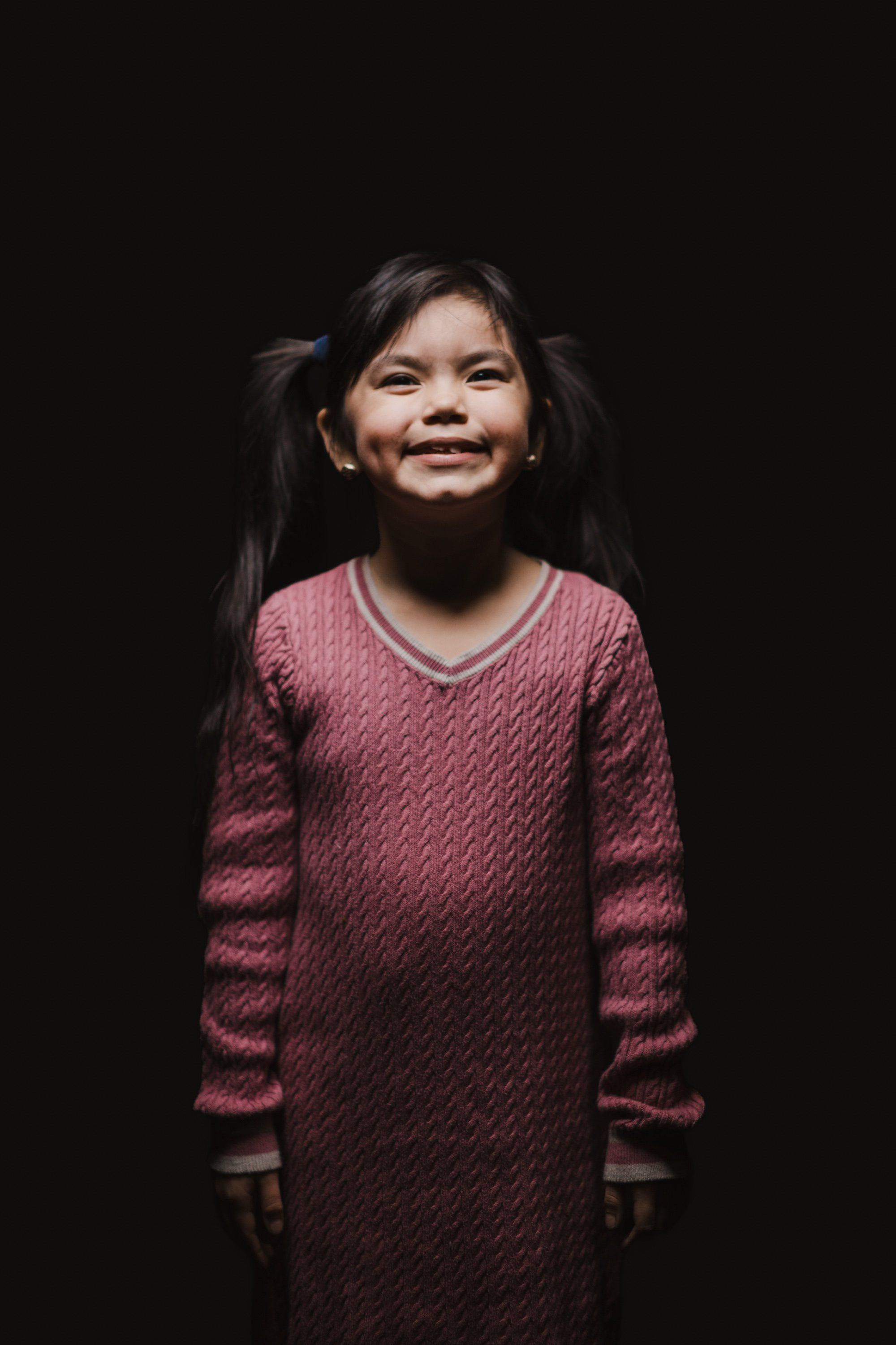 Young girl in burgundy knit dress smiling warmly against black background, long dark hair with traditional styling