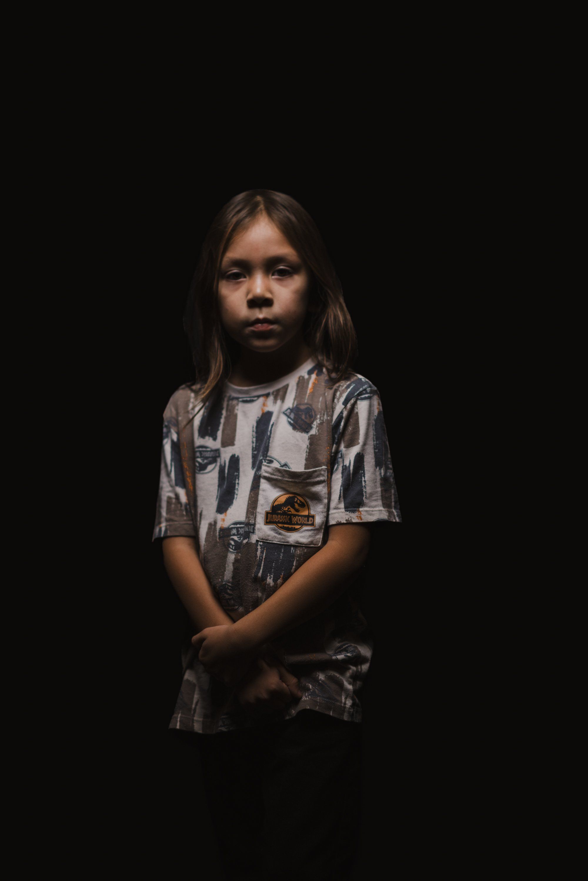 Young person in patterned gray shirt gazes solemnly against deep black background, hands clasped before them in contemplative portrait lighting