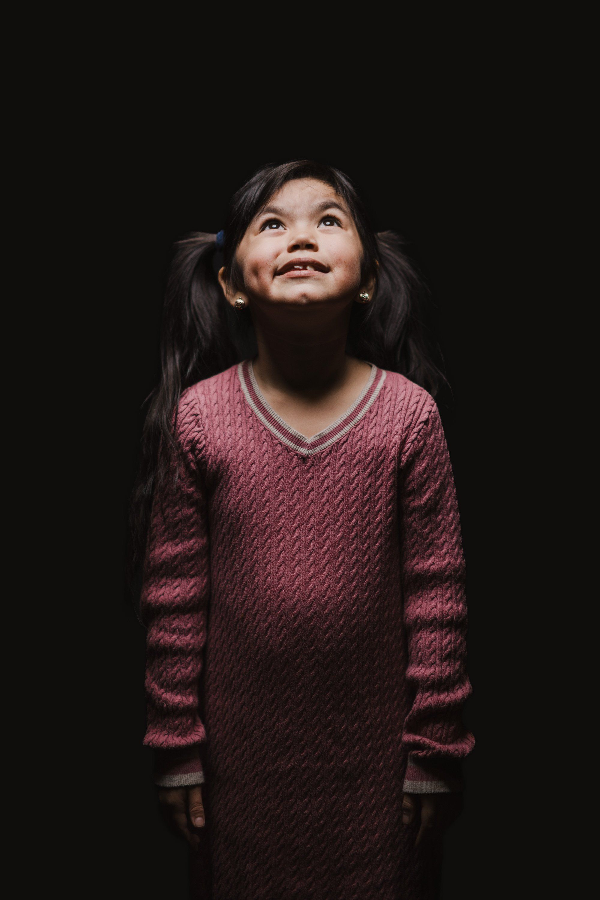 Young girl in burgundy cable-knit sweater gazes upward against dark background, expression radiant and contemplative