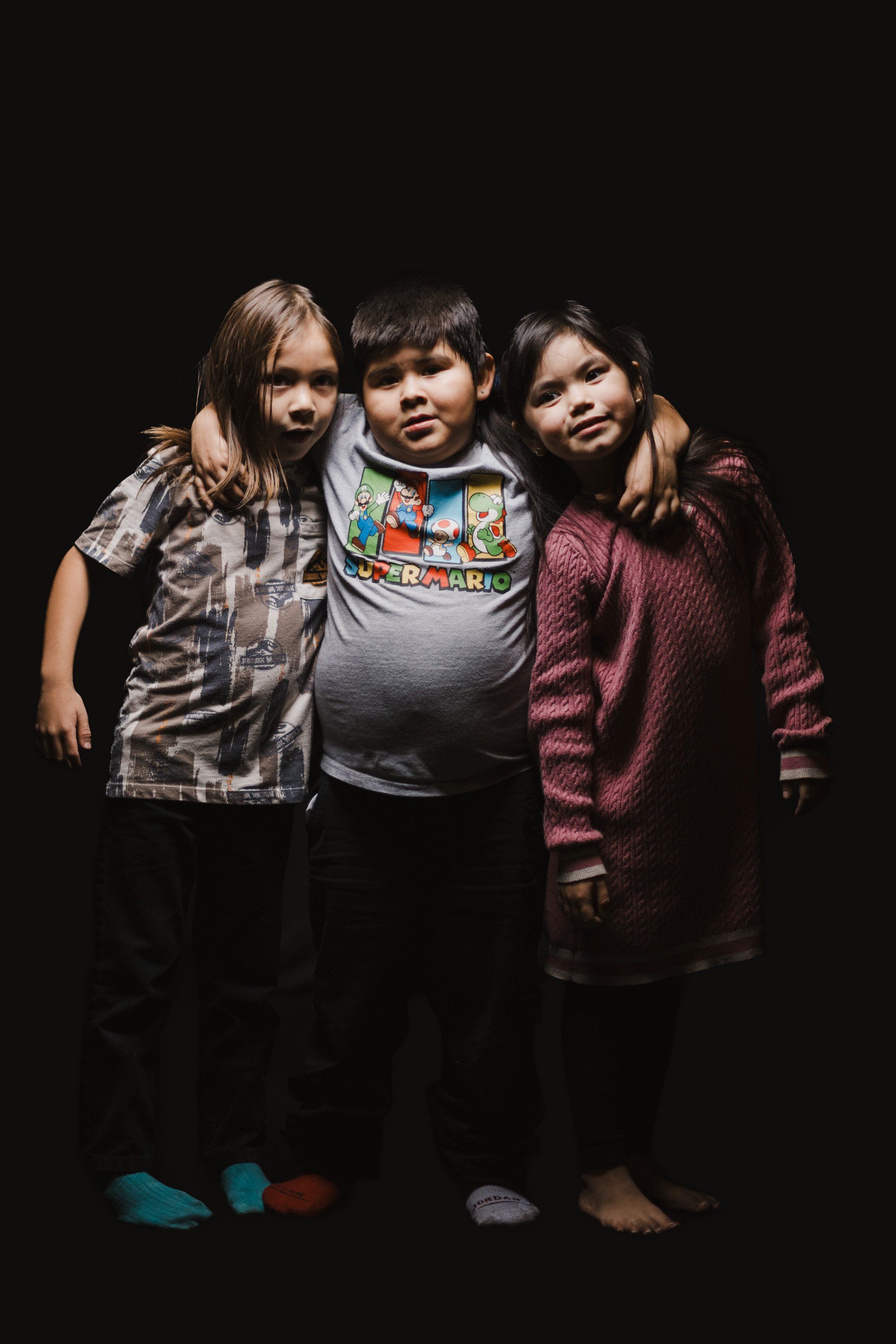 Three young children in casual clothing stand close together against a black background, warmly embracing in soft studio light
