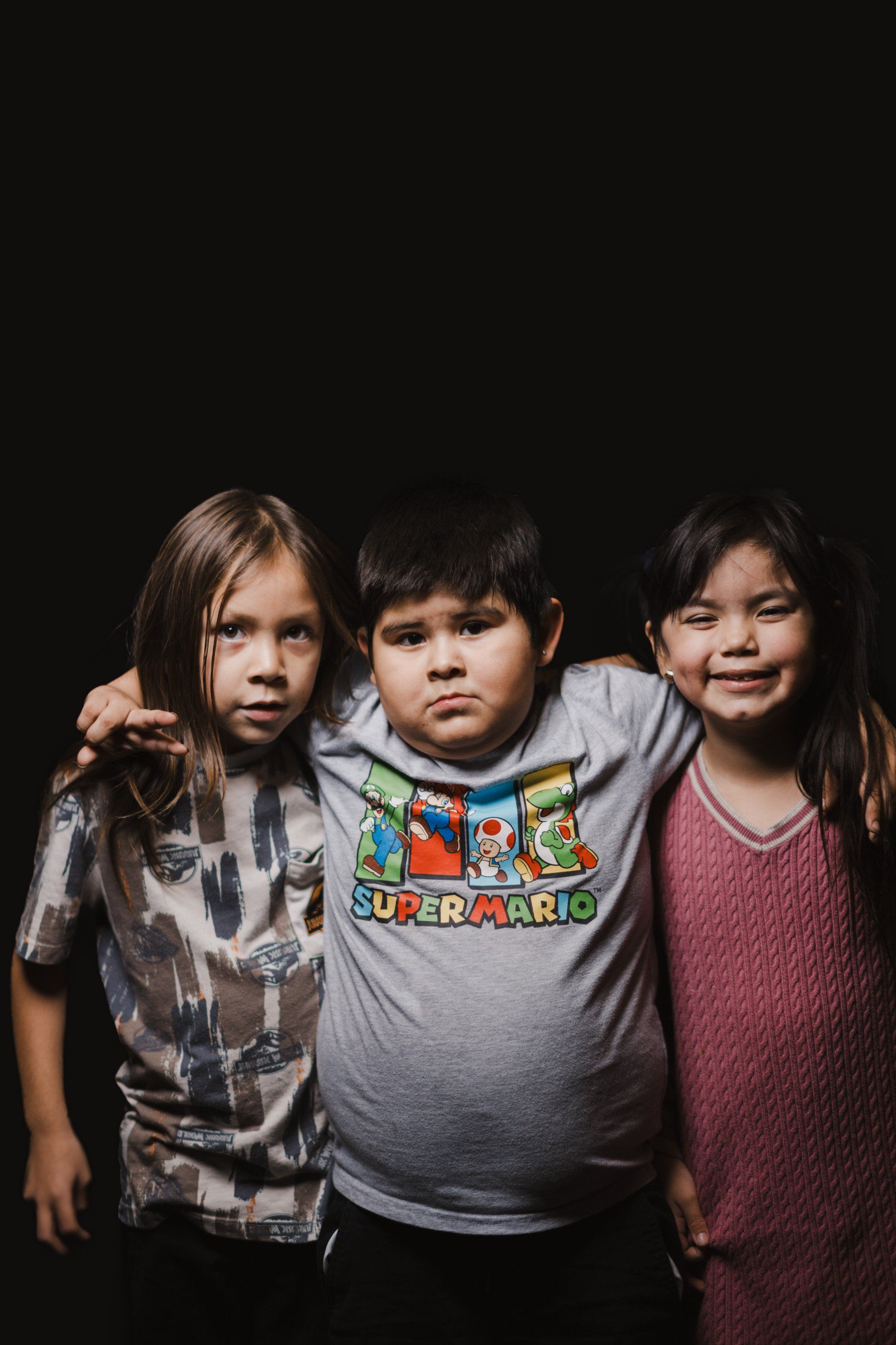 Three young people pose together against a black background, wearing casual clothing including a Super Mario graphic tee, with warm studio lighting highlight…