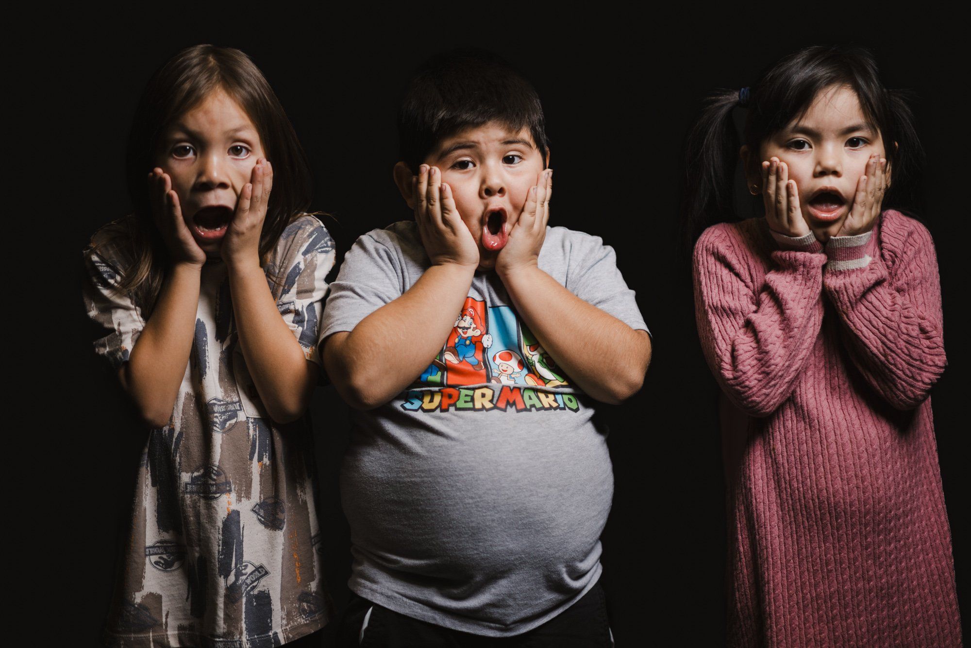 Three children with hands to their faces express surprise and delight against a dark background, celebrating together with joyful energy