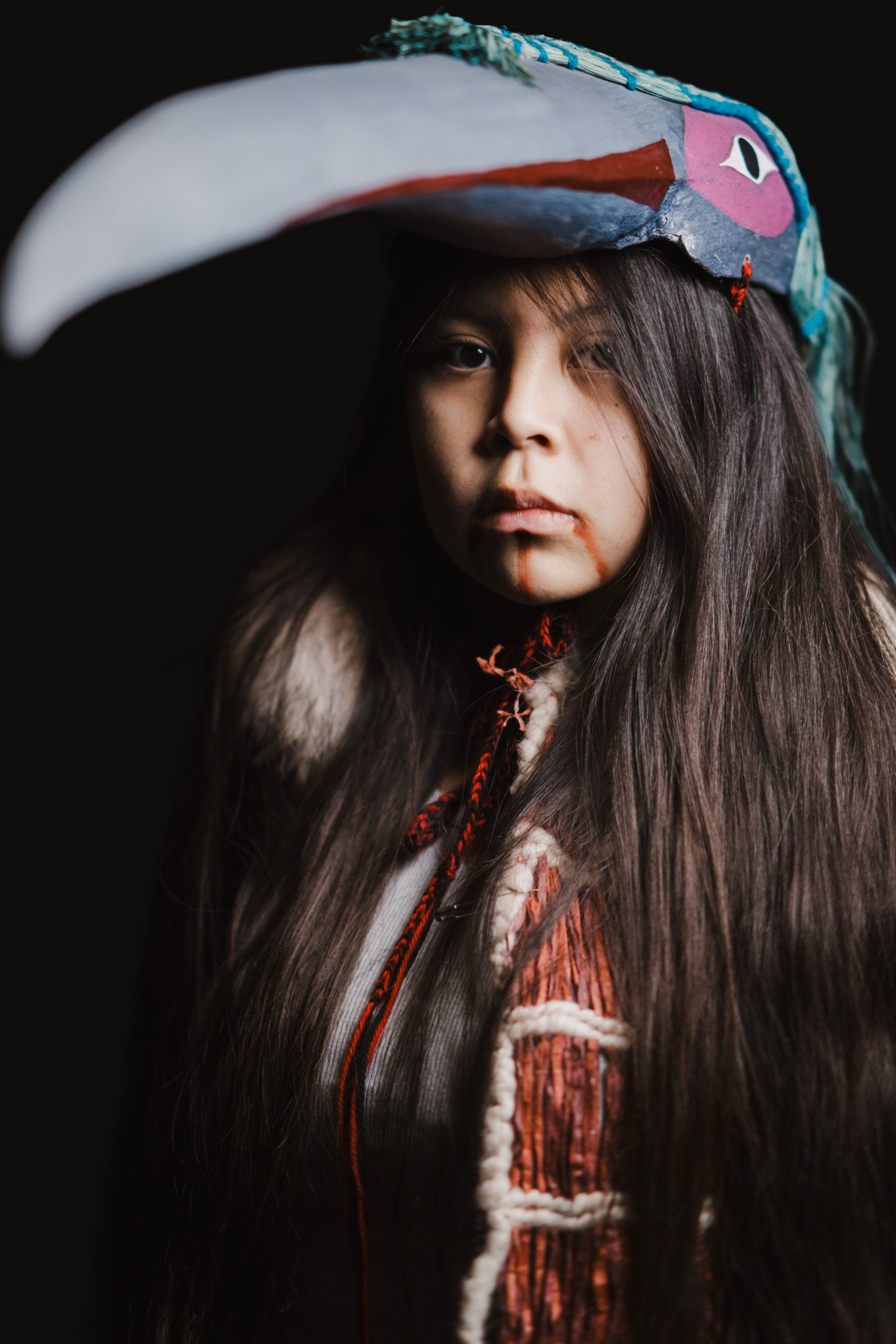 Indigenous youth in traditional regalia with long dark hair and ornate teal bird headdress against dark background, direct gaze