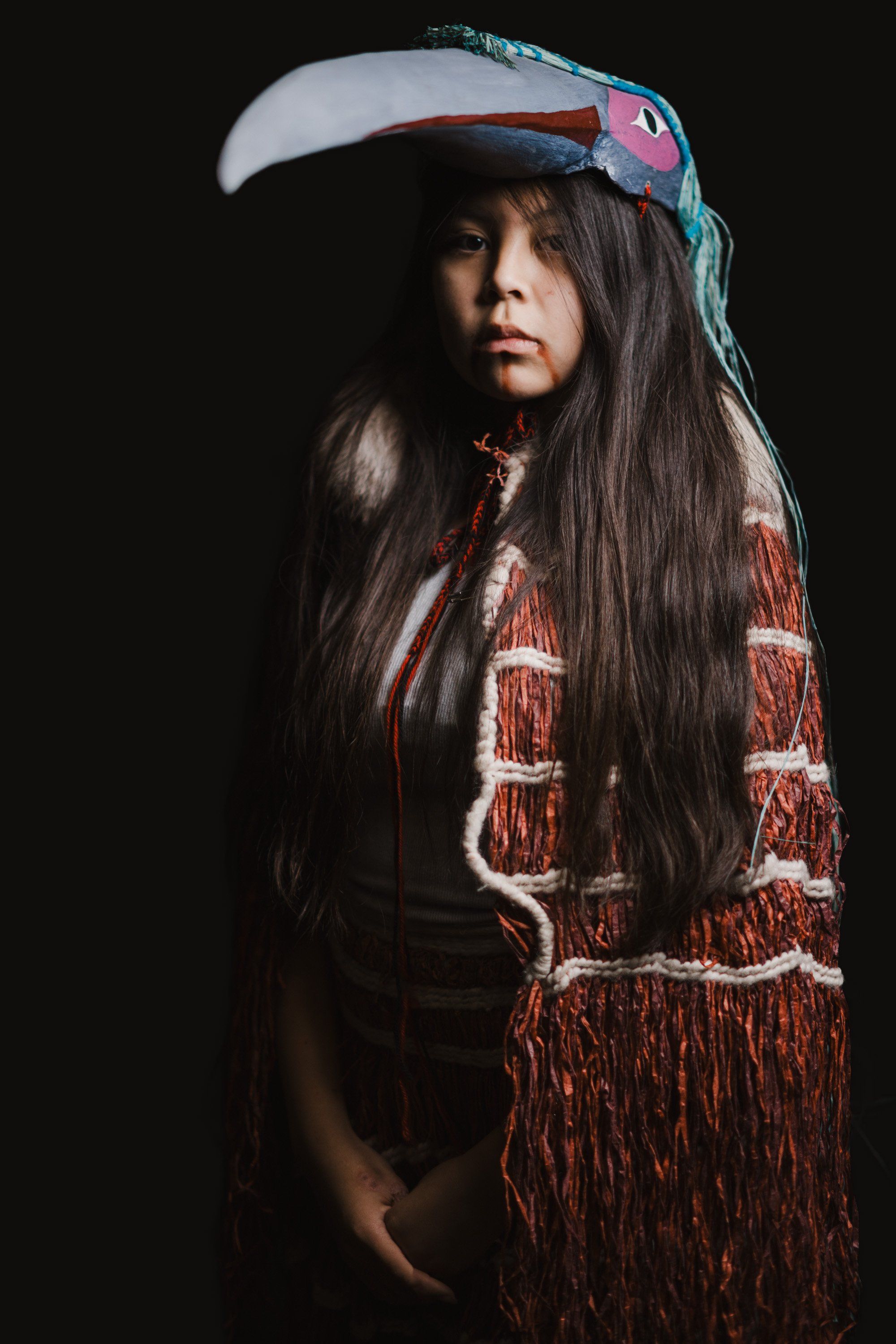 Young performer in traditional Coast Salish regalia with cedar fringe and carved mask against black background, long dark hair, contemplative gaze