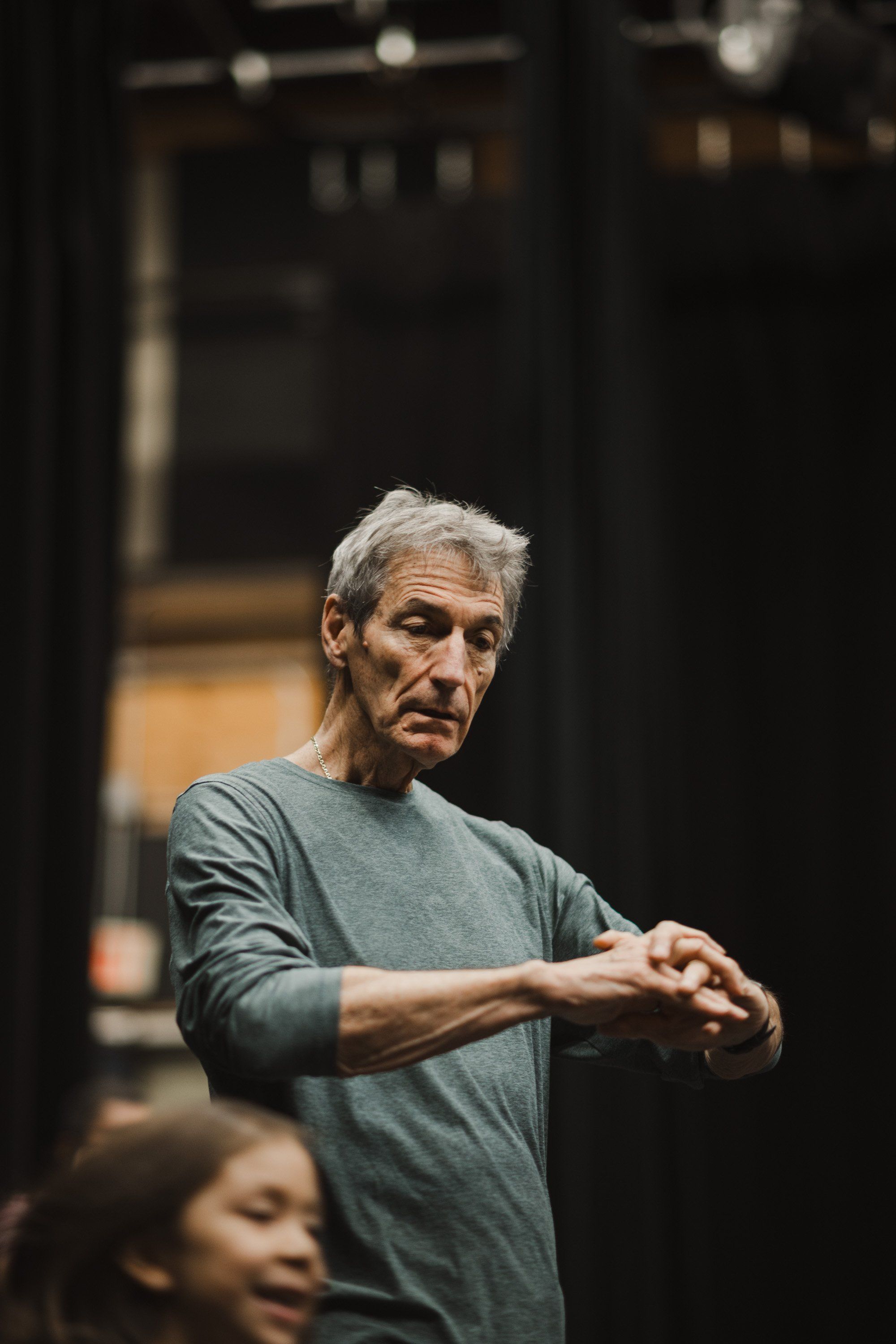 Elder in grey t-shirt gestures mid-movement during Coast Salish performance, warm stage lighting illuminating focused expression