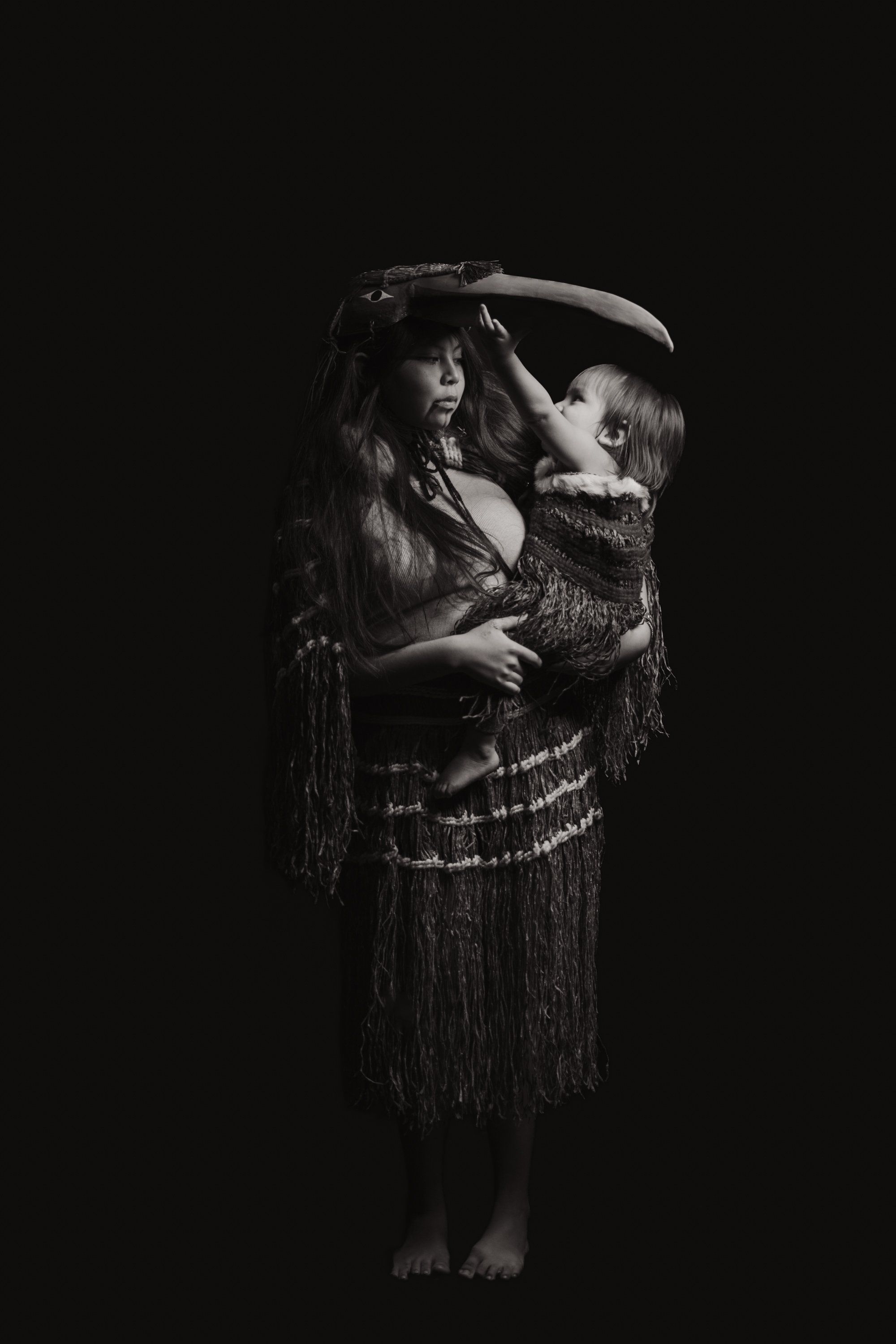 Child in traditional regalia with fringed blanket held aloft by adult against black background, intimate moment of cultural transmission