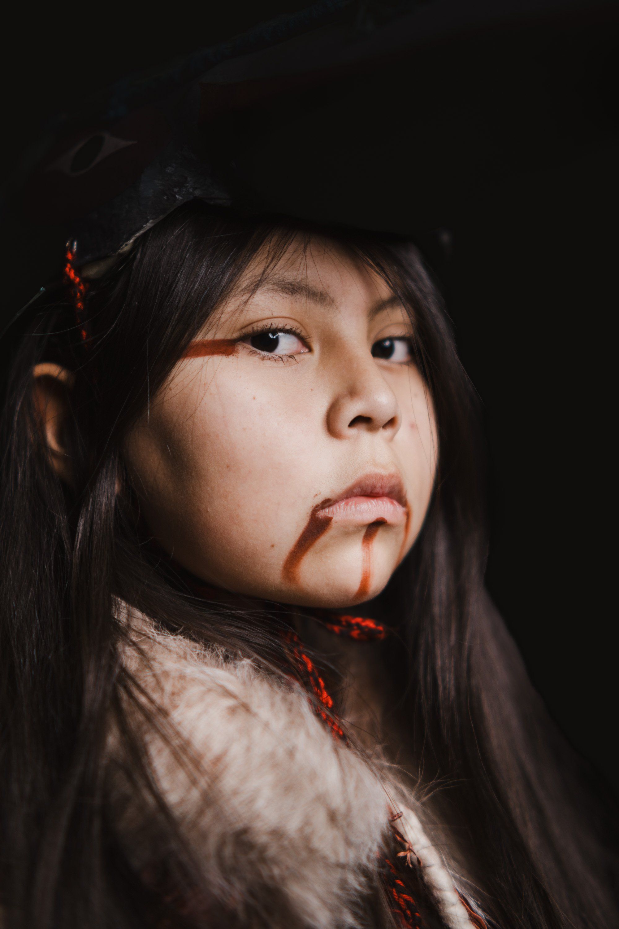 Young person with long dark hair and red face paint against black background, gazing upward with contemplative expression