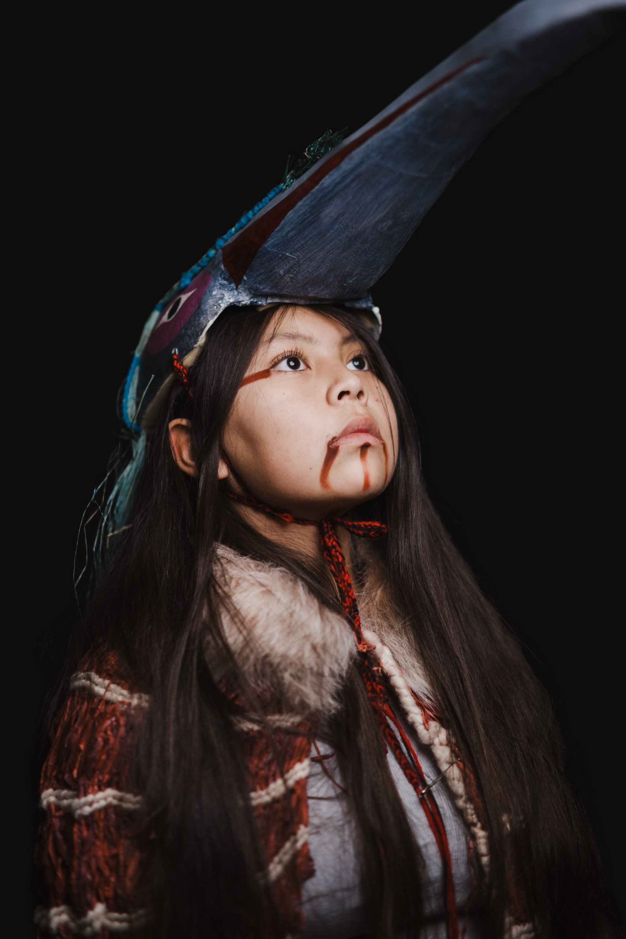 Young dancer in traditional regalia looks upward, face painted with red designs, large feathered headdress silhouetted against black background