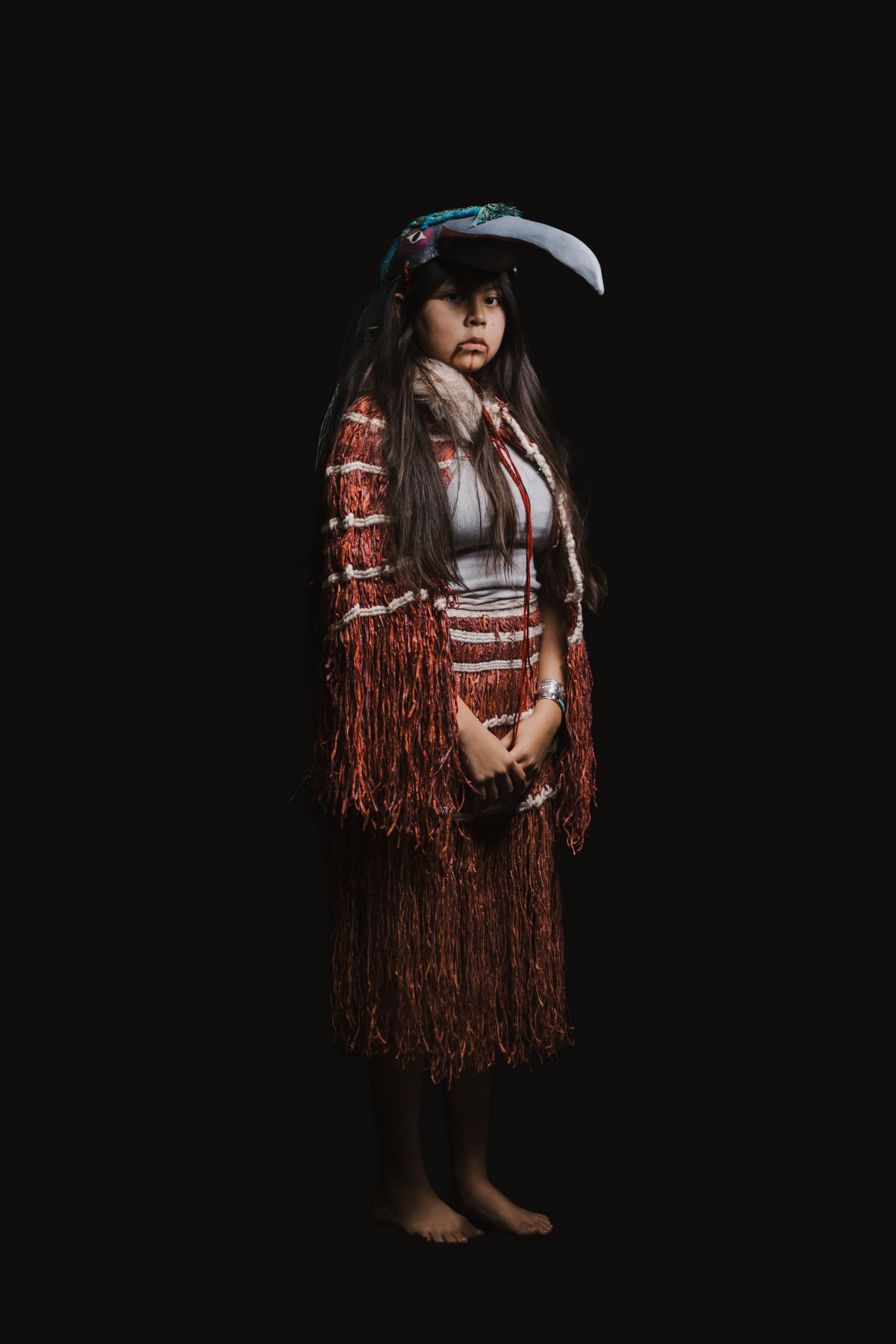 Young dancer in traditional Coast Salish regalia with fringed cedar skirt and button blanket stands against black background
