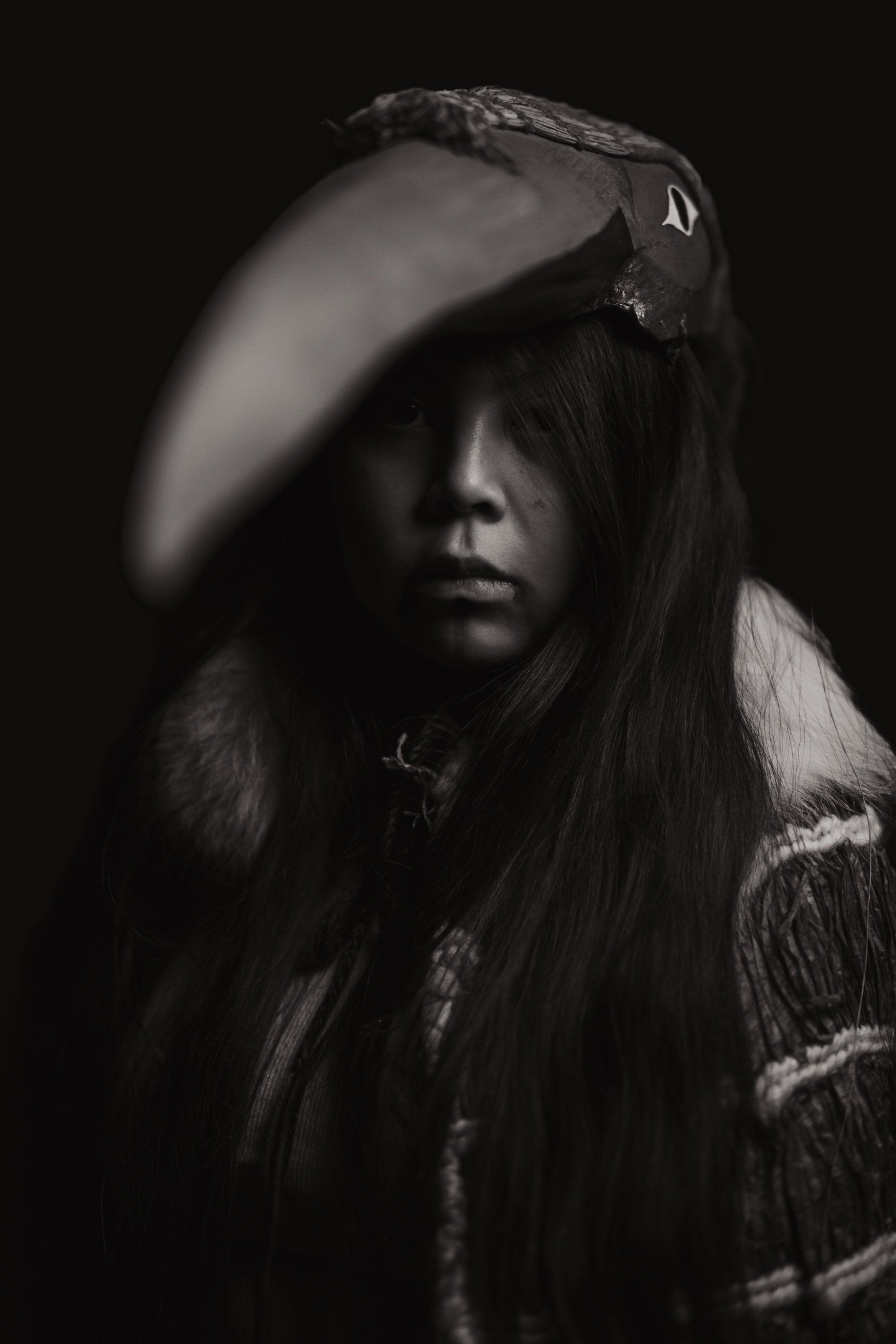 Portrait of a young person with long dark hair wearing traditional regalia including a carved bird headdress, photographed against black with dramatic side l…