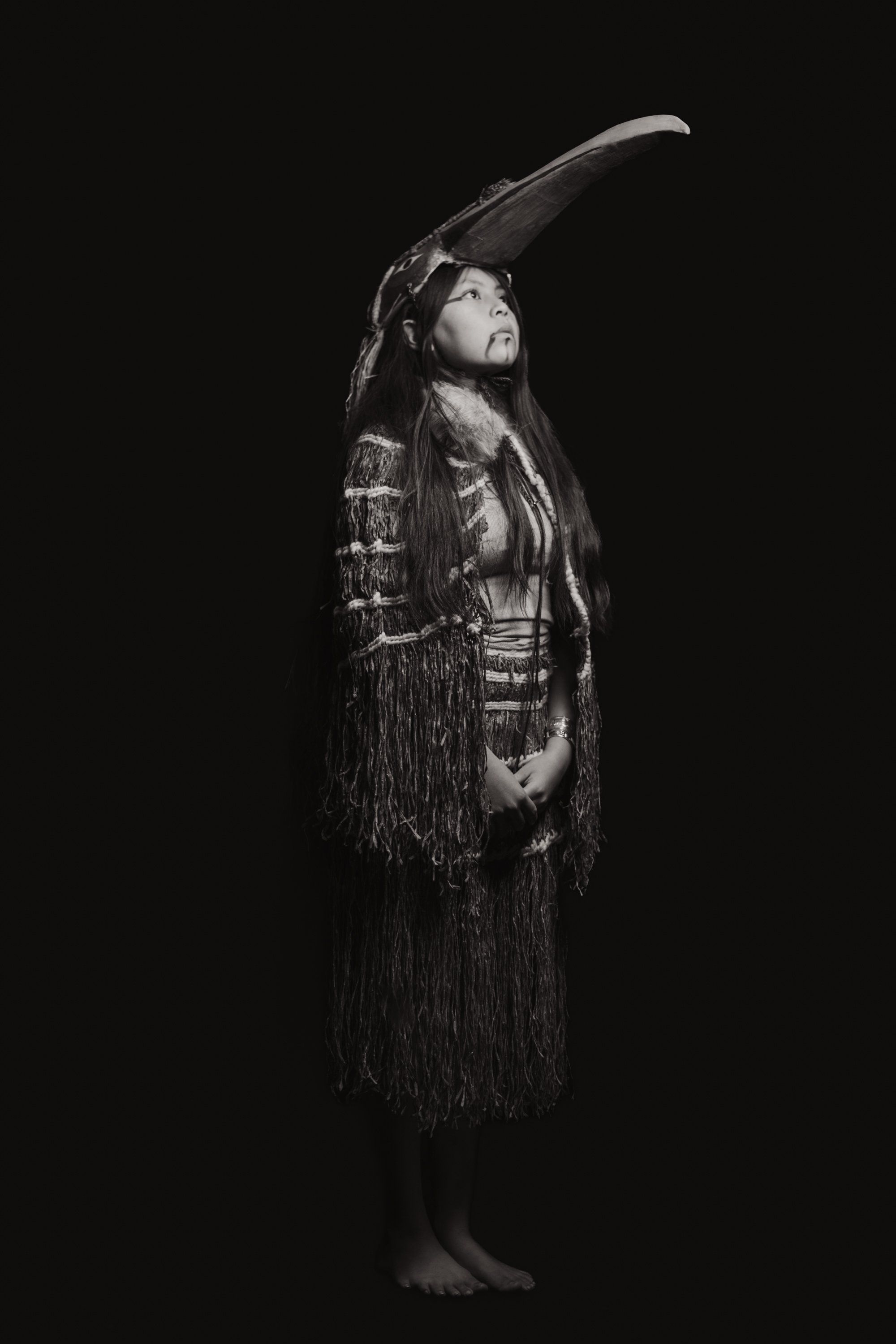 Woman in traditional Coast Salish regalia with fringed shawl and horn headdress gazes upward against black background