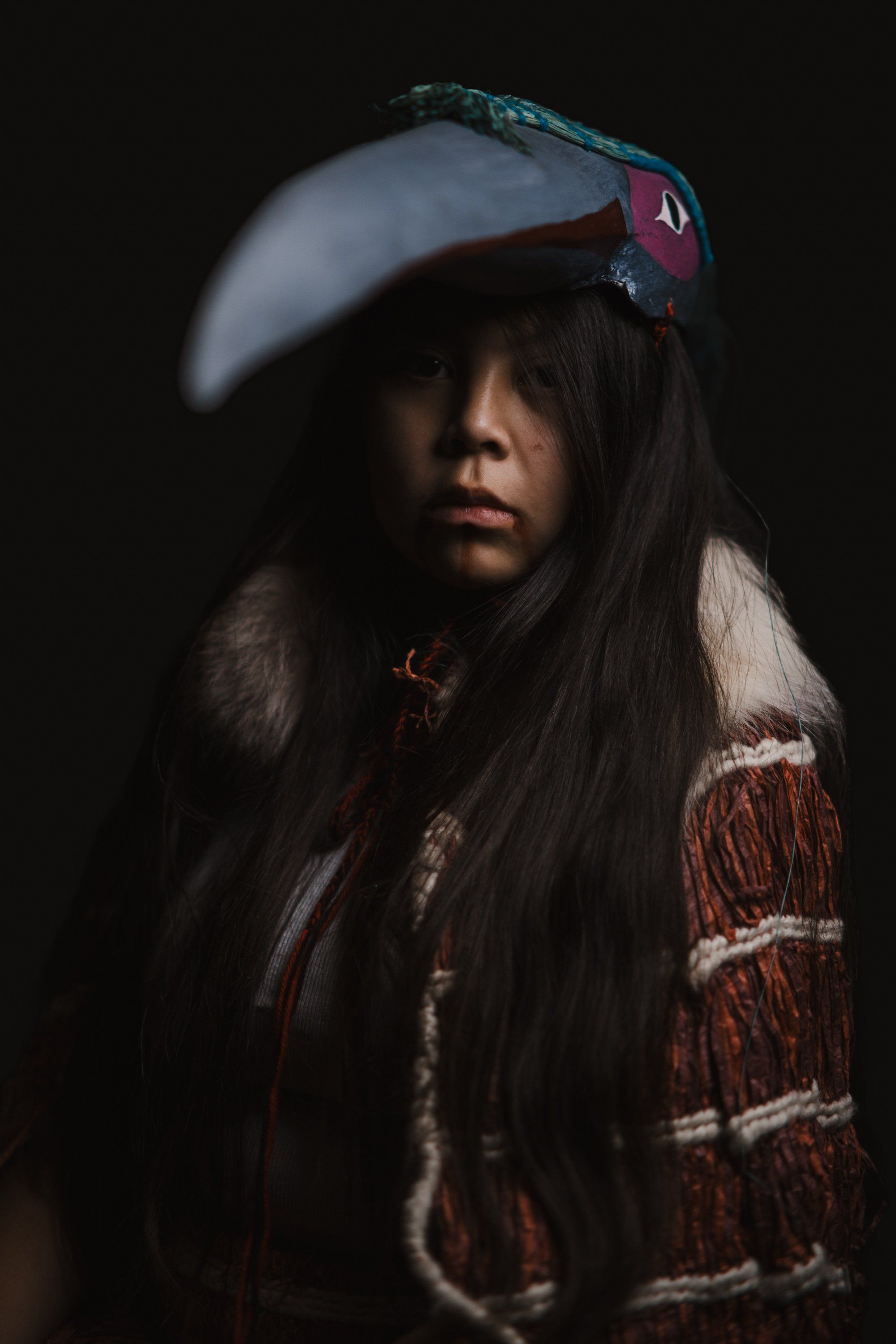 Young person in traditional Coast Salish regalia with long dark hair, wearing a decorated button blanket and feathered headdress, posed against dark backgrou…