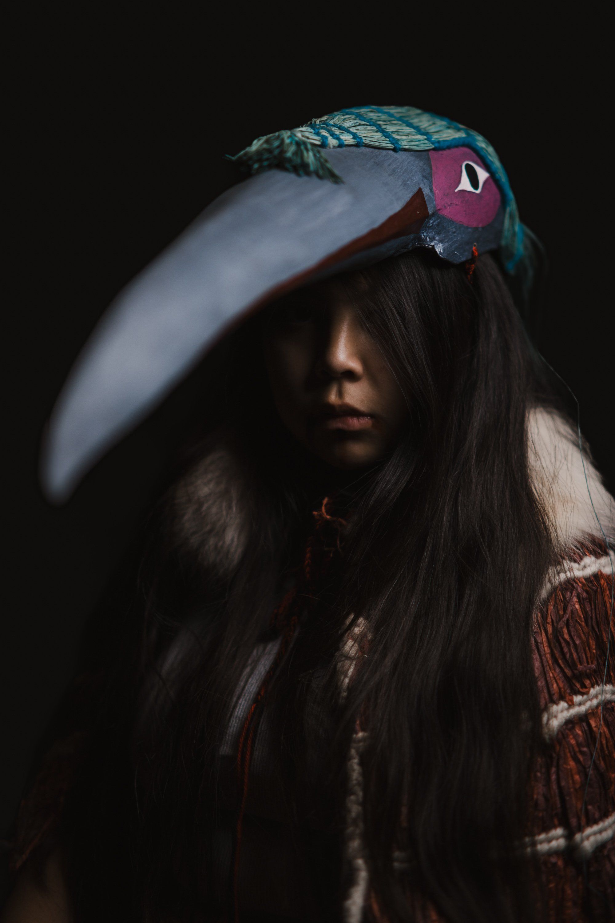 Person with long dark hair wearing an ornate teal and pink bird headdress against a black background, dramatic side lighting
