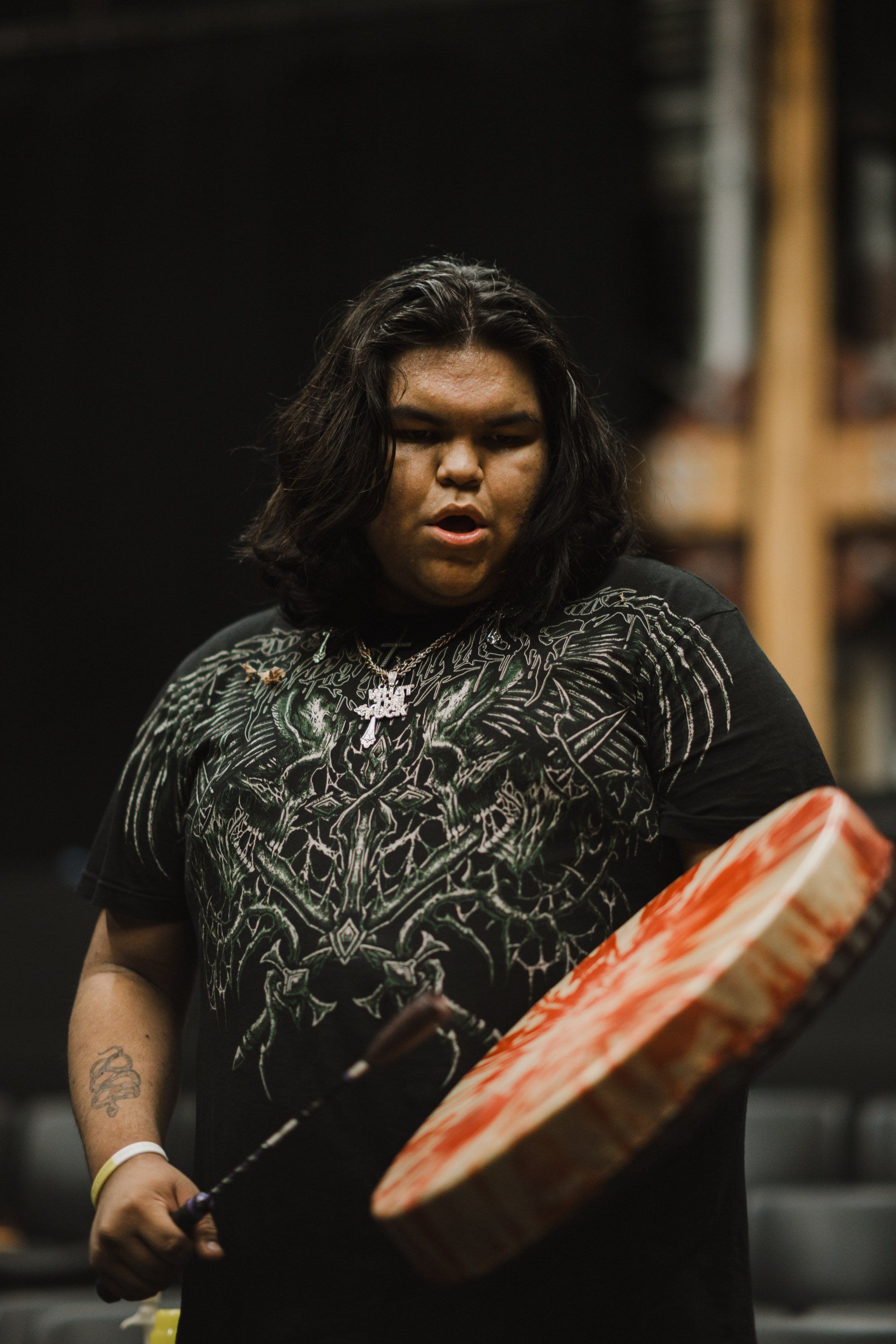 Performer in ornate black graphic tee holds a carved wooden drum against dark indoor backdrop, focused expression captured in warm stage lighting
