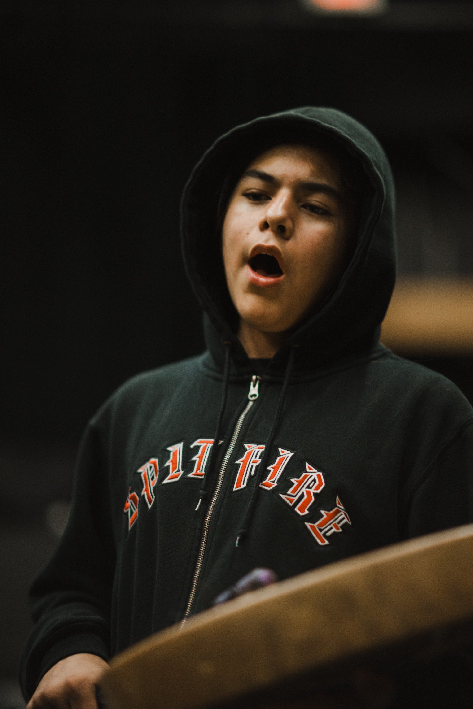 Young performer in black hoodie with text detail, mouth open mid-song, warm stage lighting against dark background