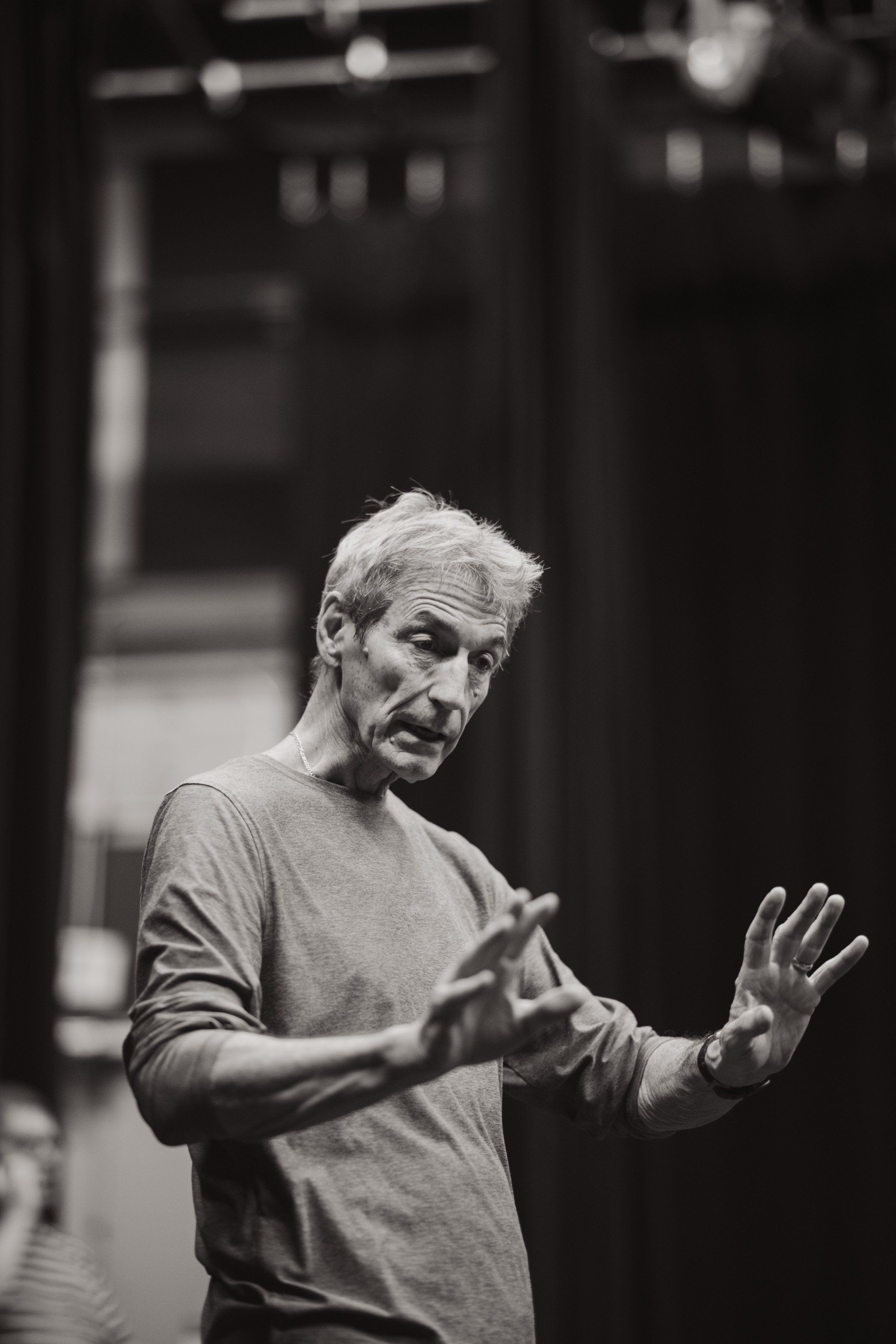 Elder gestures expressively while speaking, hands raised in animated conversation, black and white portrait against dark interior backdrop