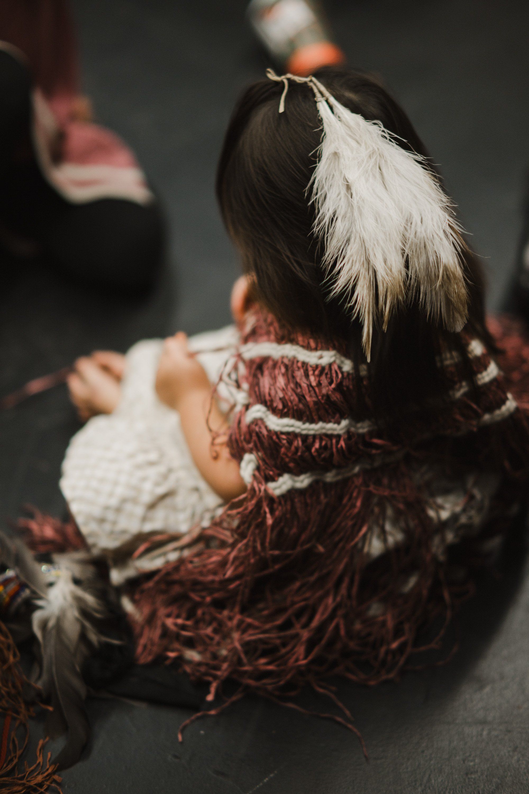 Child from behind wearing burgundy knit cardigan and white feather hair ornament, soft natural light