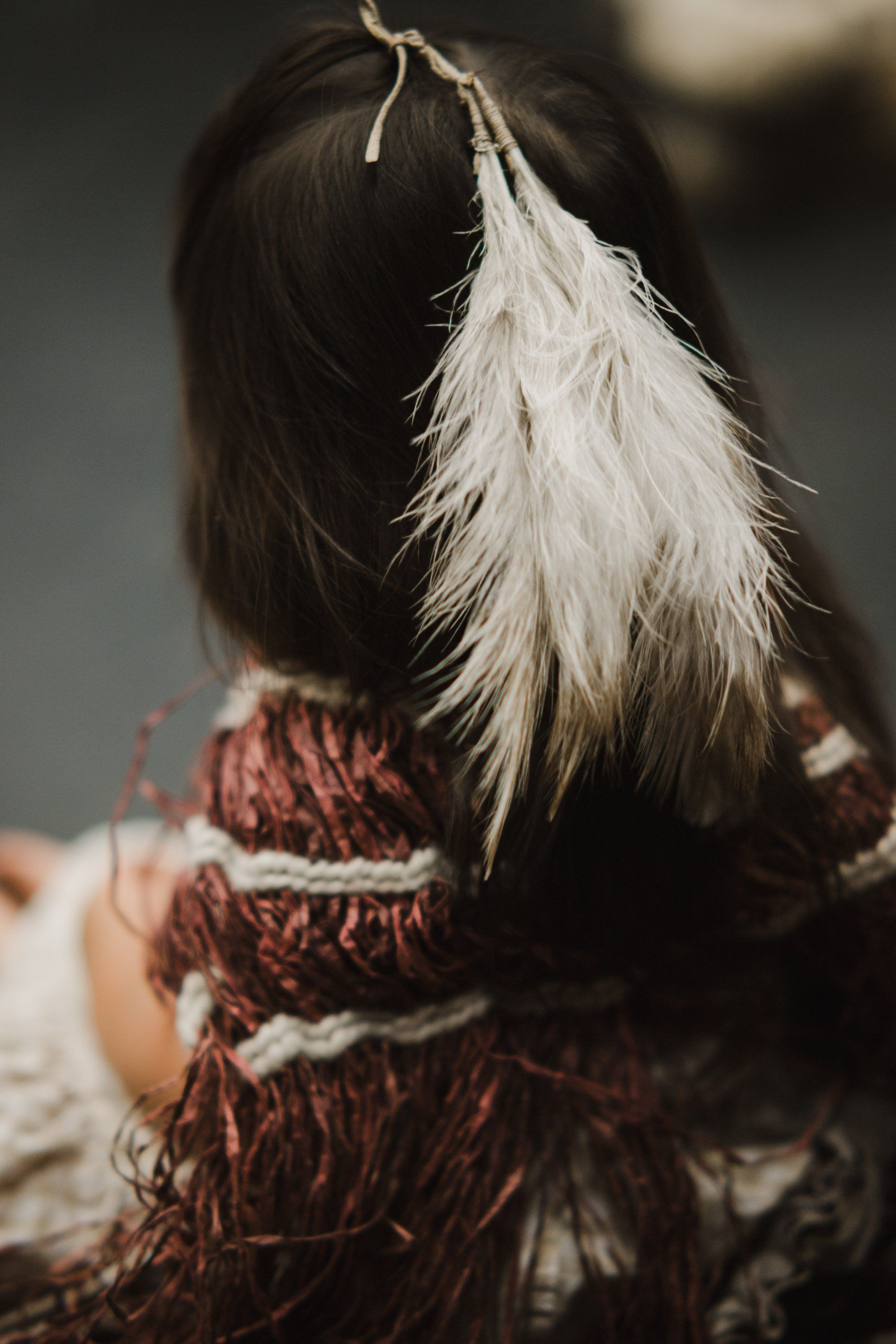 Dark-haired person wearing rust-red regalia viewed from behind, white feather hair ornament catching soft light against blurred background
