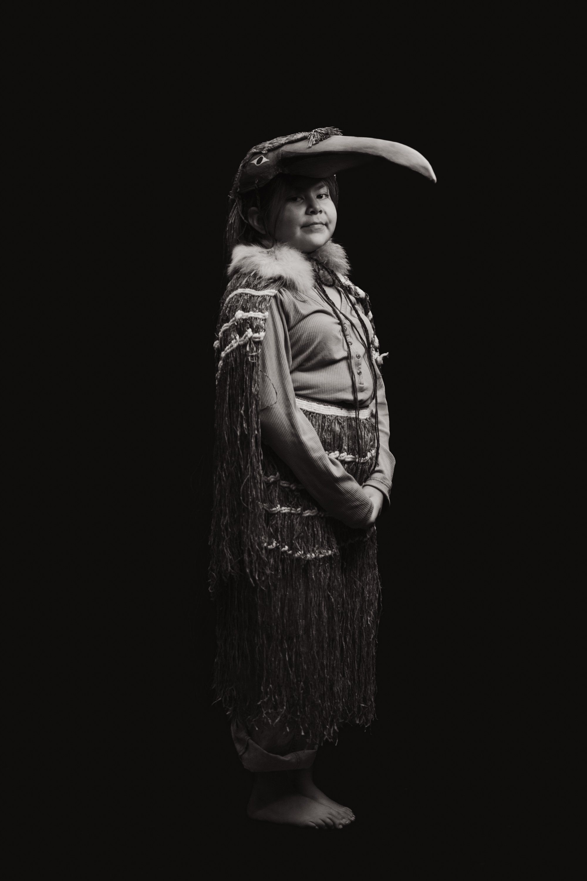 Portrait of a dancer in traditional Coast Salish regalia, including a curved hat and fringed cedar clothing, posed against black background