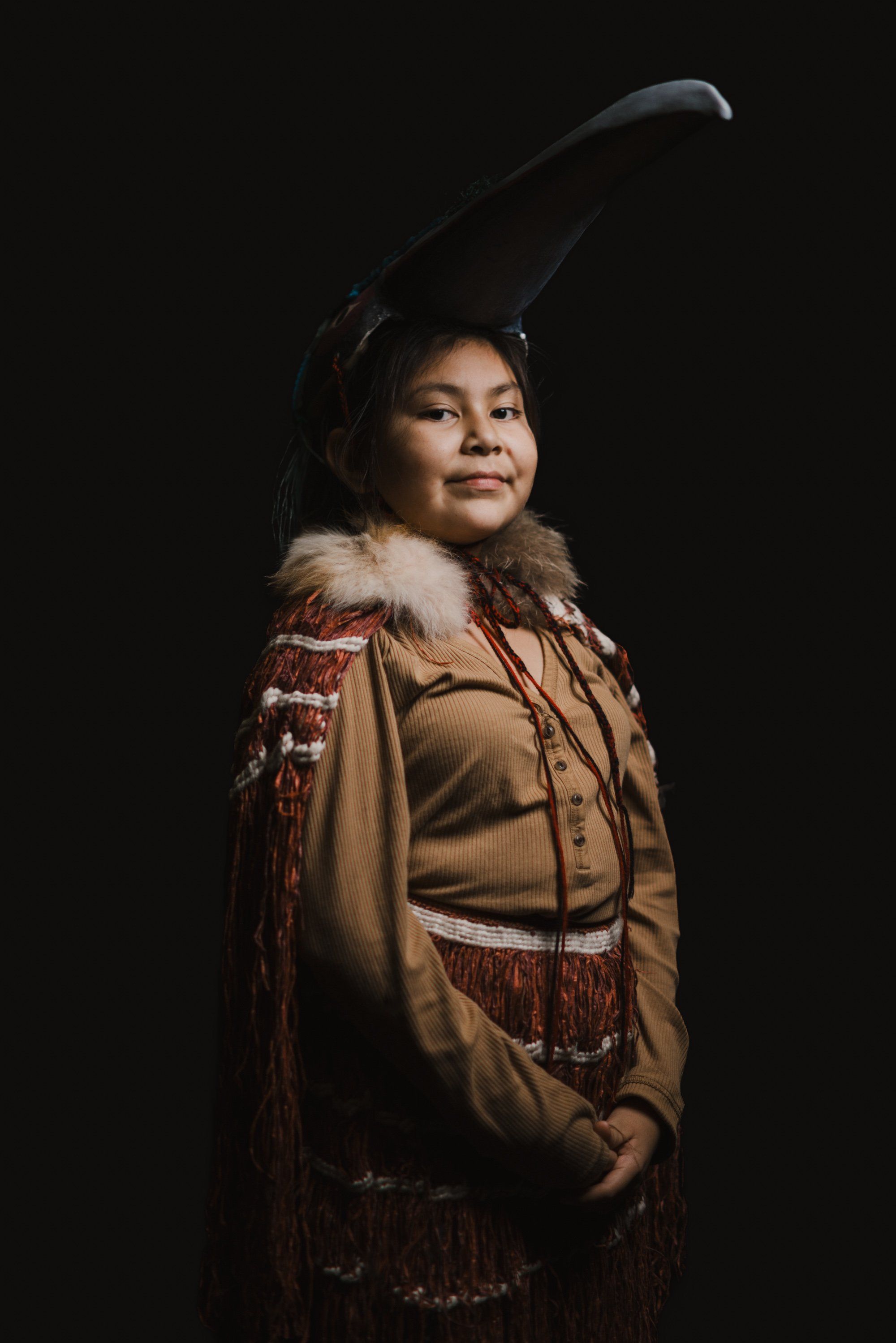 Young person in traditional Coast Salish regalia with tall feathered headdress, fur-trimmed tan tunic, and beaded details, posed against black background wit…