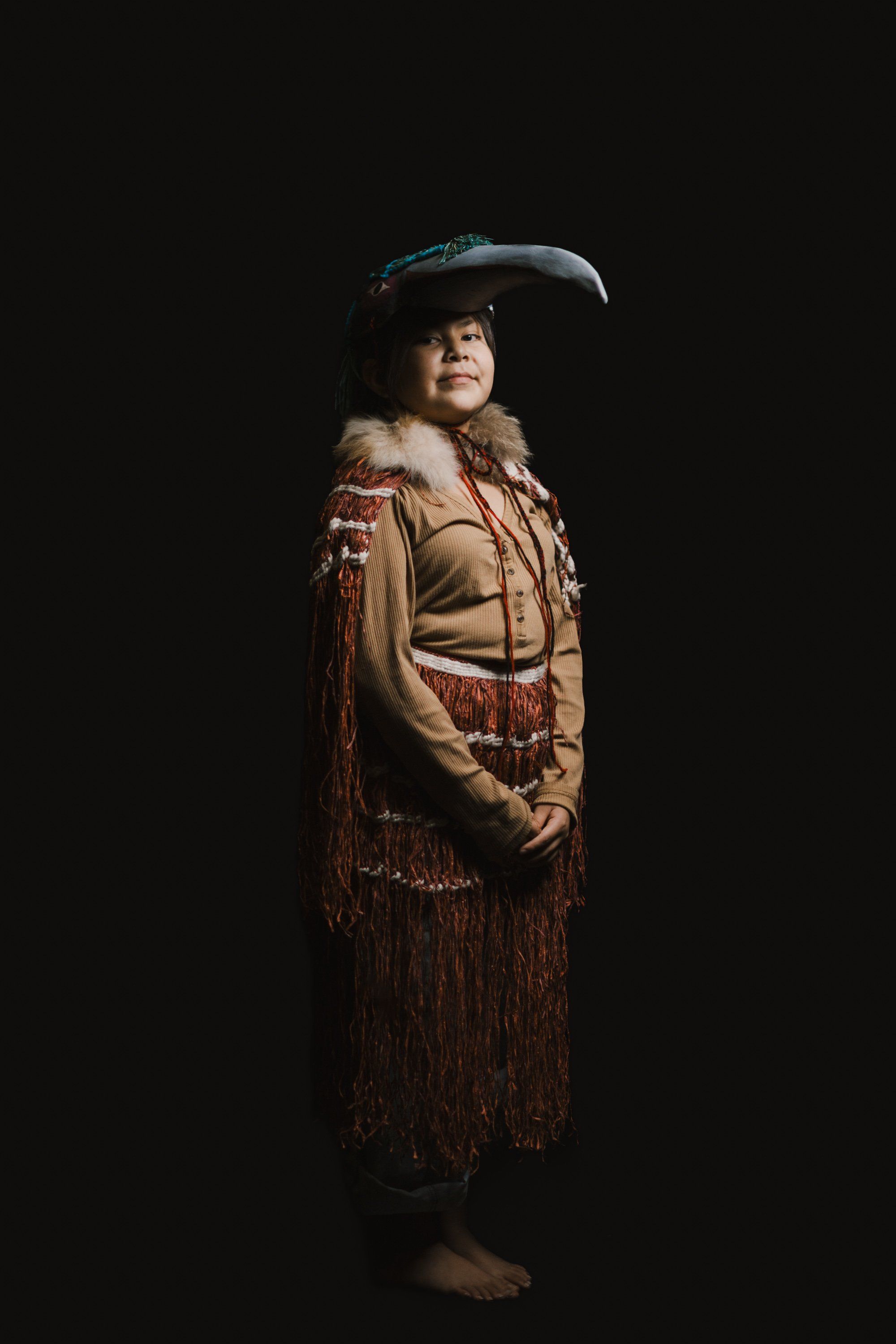 Coast Salish dancer in traditional regalia with fur-trimmed button blanket and carved hat, posed against black background with dramatic side lighting