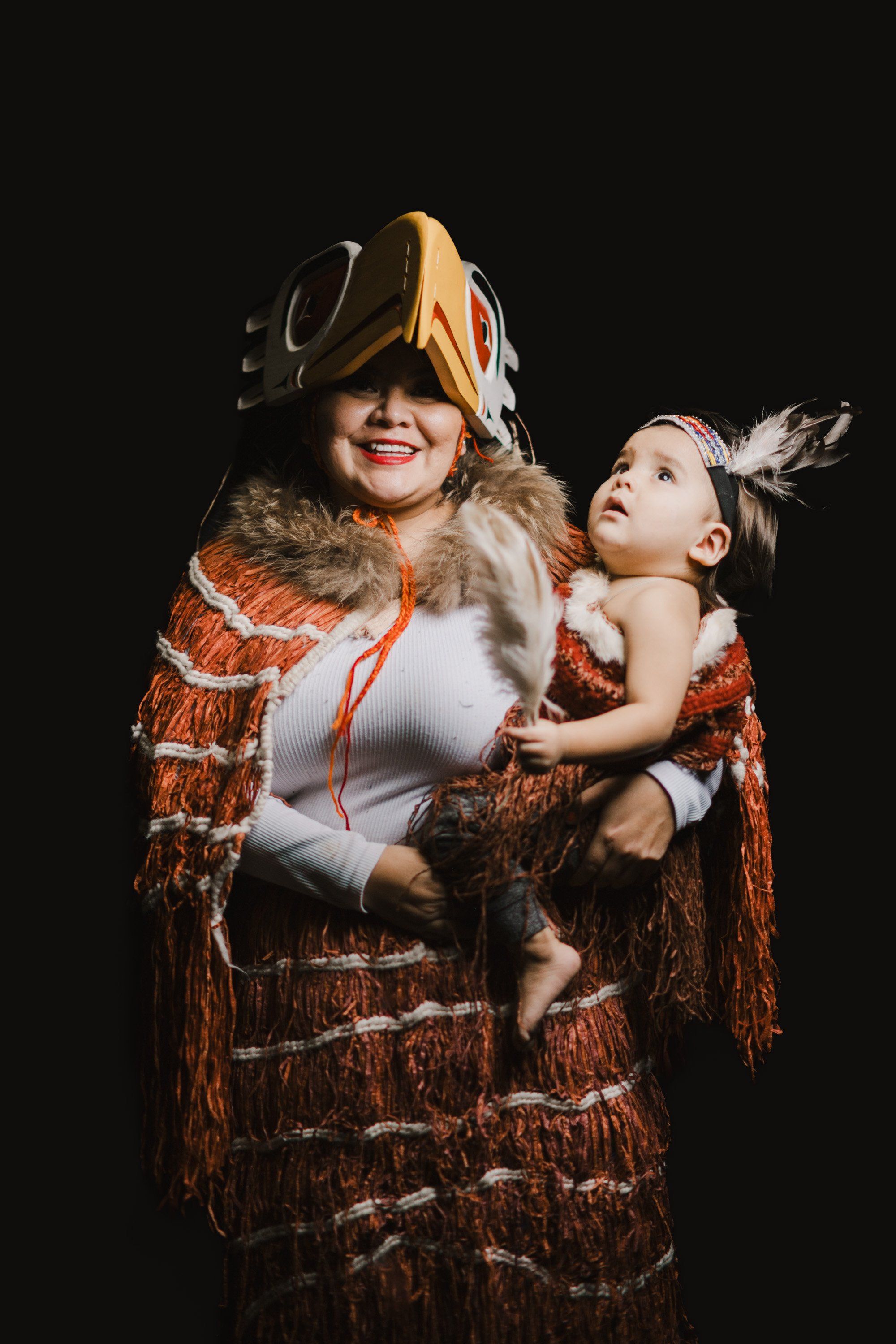 Adult and child in rust-patterned regalia with fur trim and feathered headdresses embrace against black background, their joyful expressions celebrating Coas…