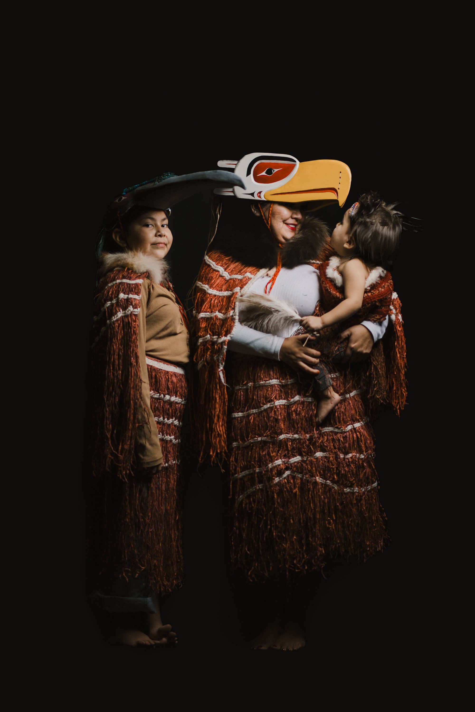 Three people in traditional Coast Salish regalia and a carved raven mask pose together against black, warmly lit from the side