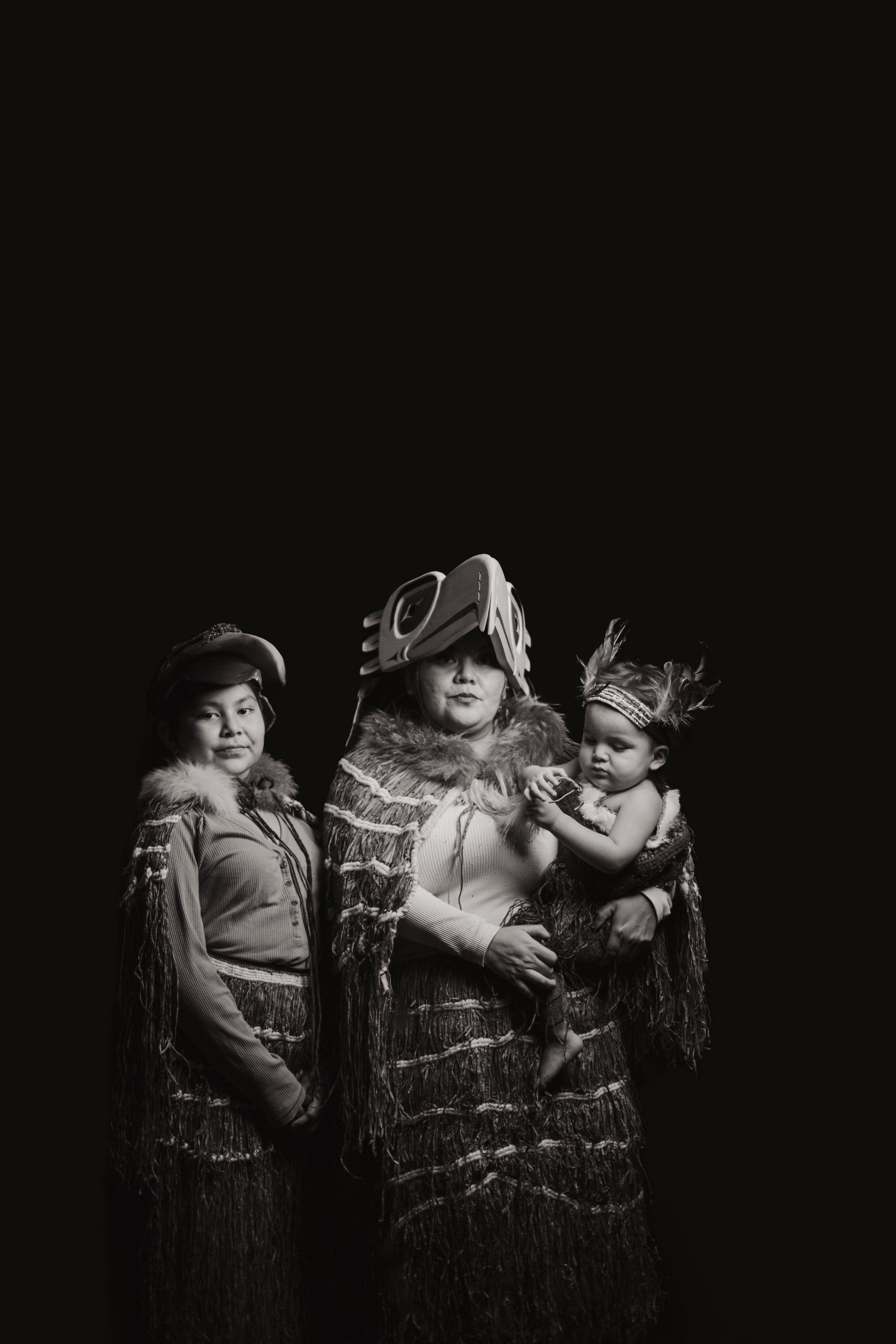 Three figures in traditional regalia with fringed clothing and headdresses pose together against black, lit dramatically in monochrome