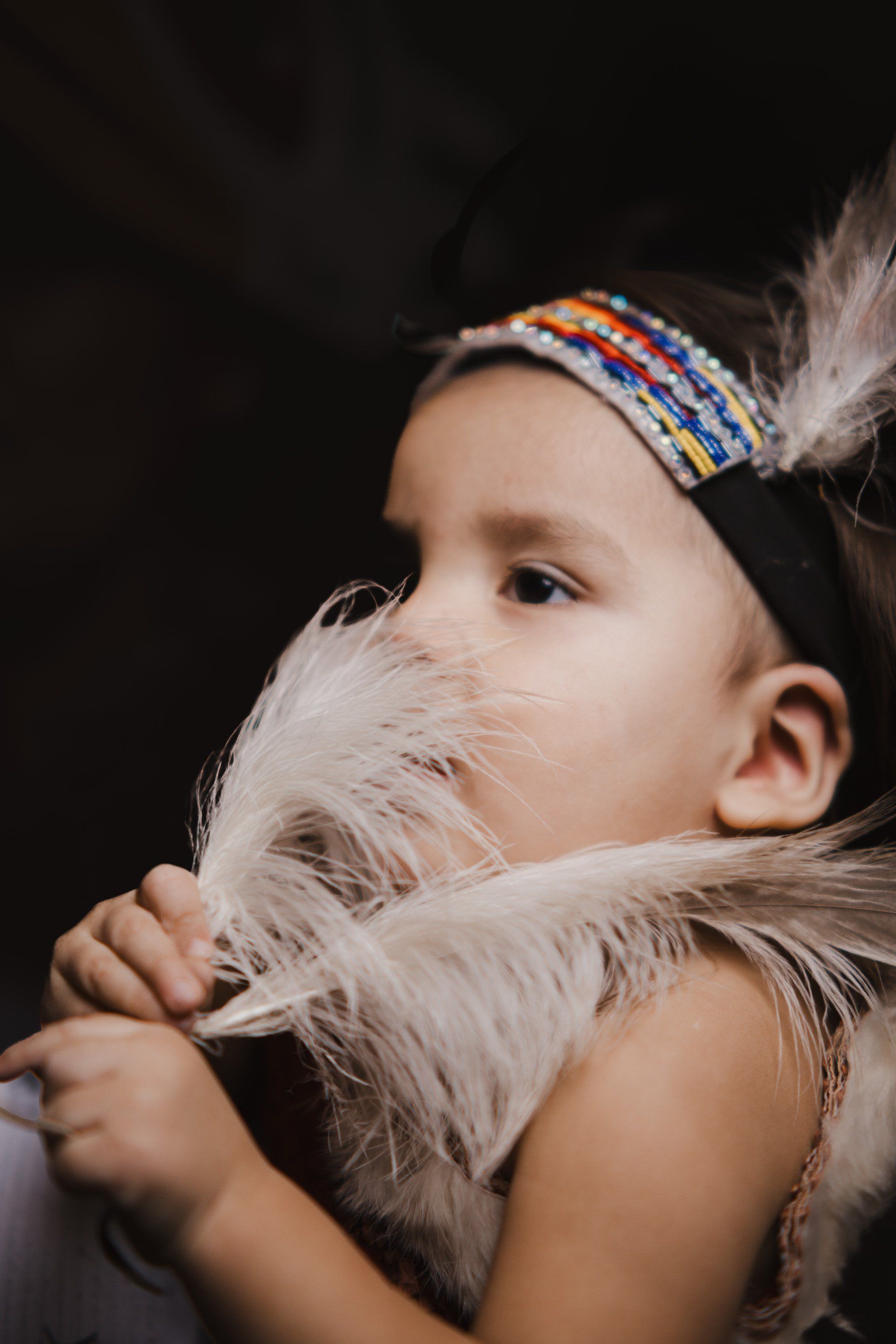 Young child holding a white feather near their face, wearing a beaded headband against a dark background